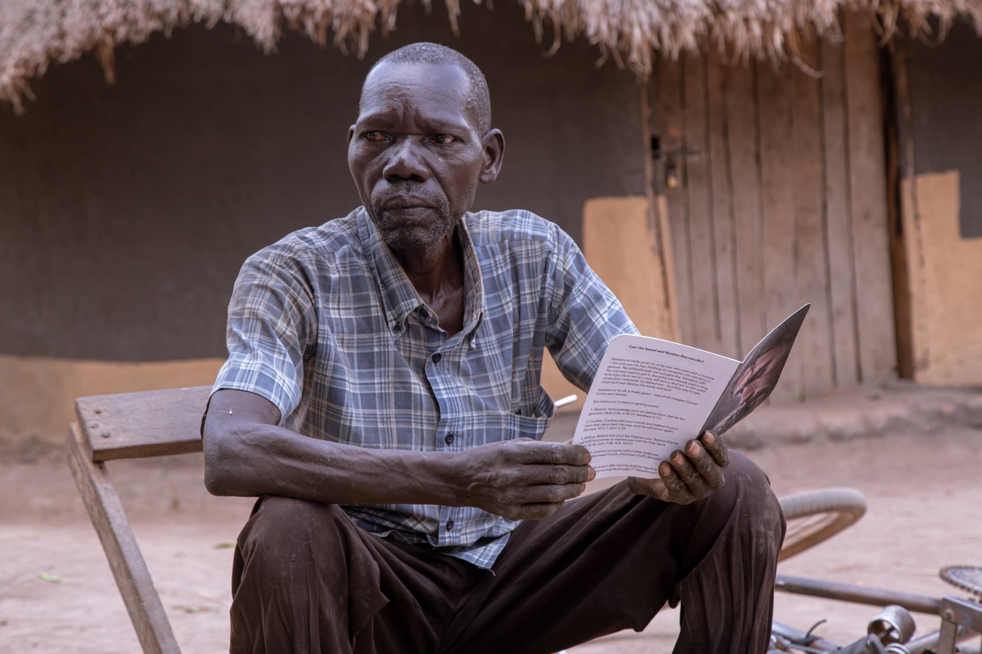 An elderly man sits outside a thatched-roof home holding folded papers and a photo.