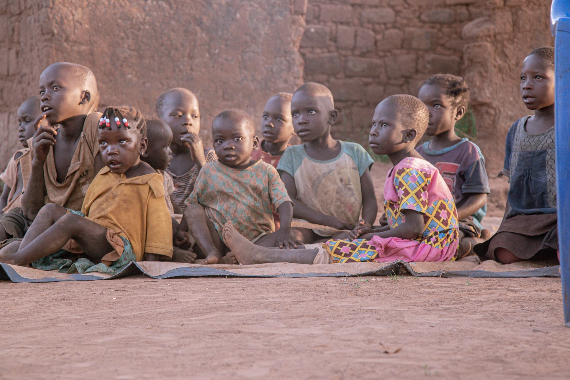 Row of children sitting together on a mat against a curved mud wall.
