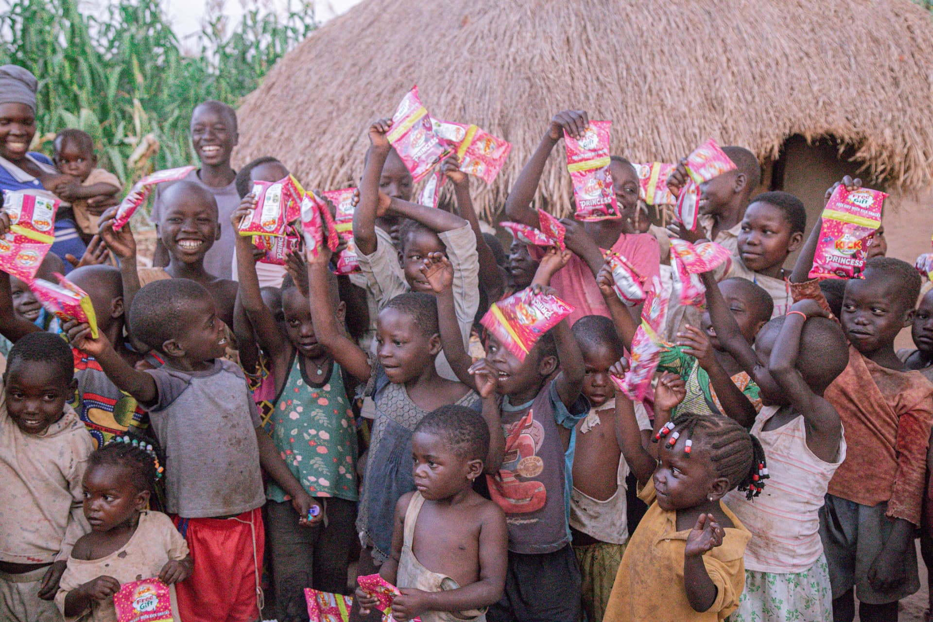 Large group of children under a thatched shelter raise pink food packets above their heads.