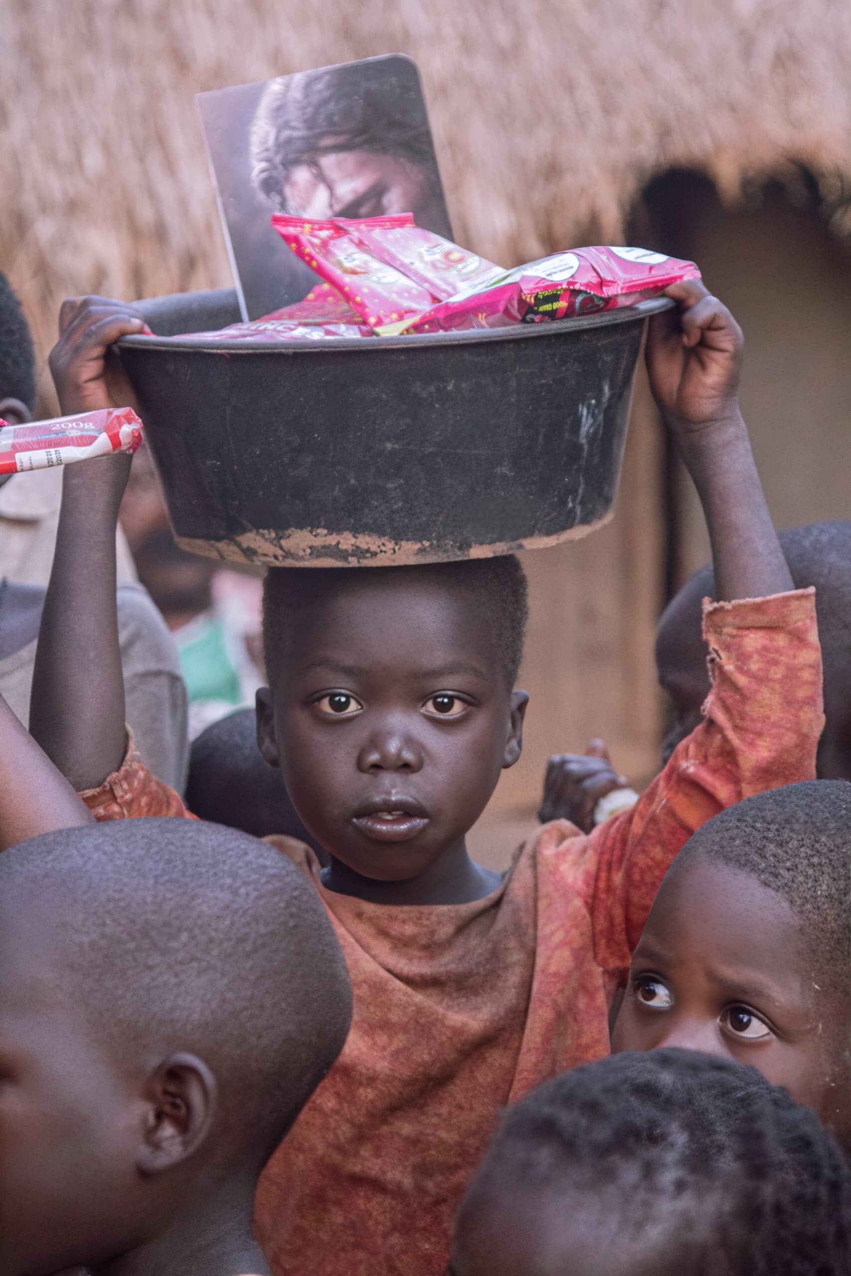 A boy balances a basin of food packets on his head, surrounded by other children.