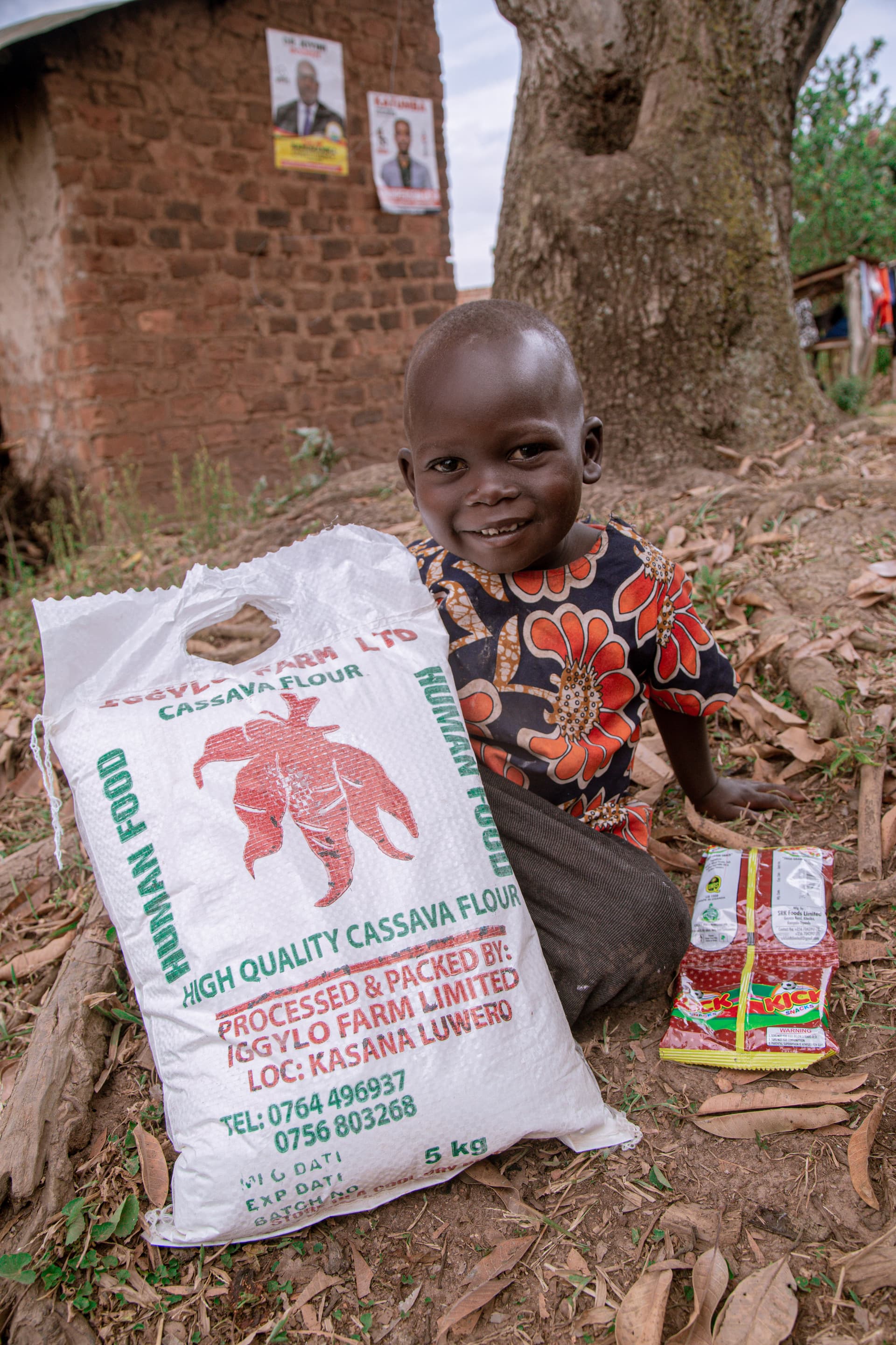 A boy stands holding a large bag of cassava flour near a brick wall.