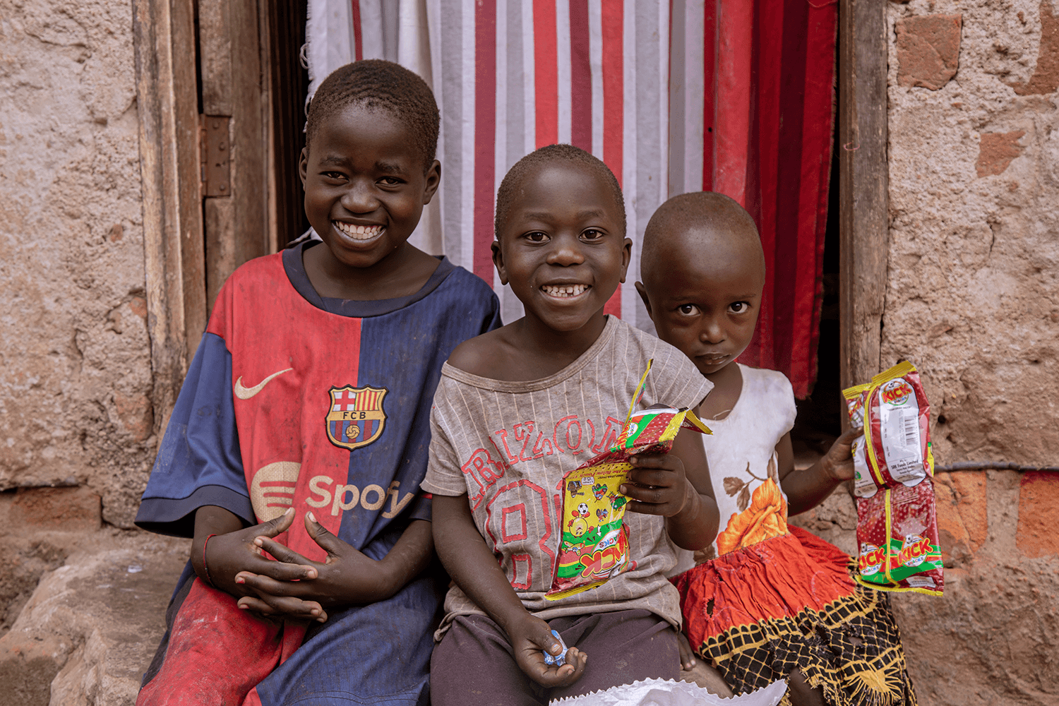 Three children sit together on a bench holding small food packets, a red curtain behind them.