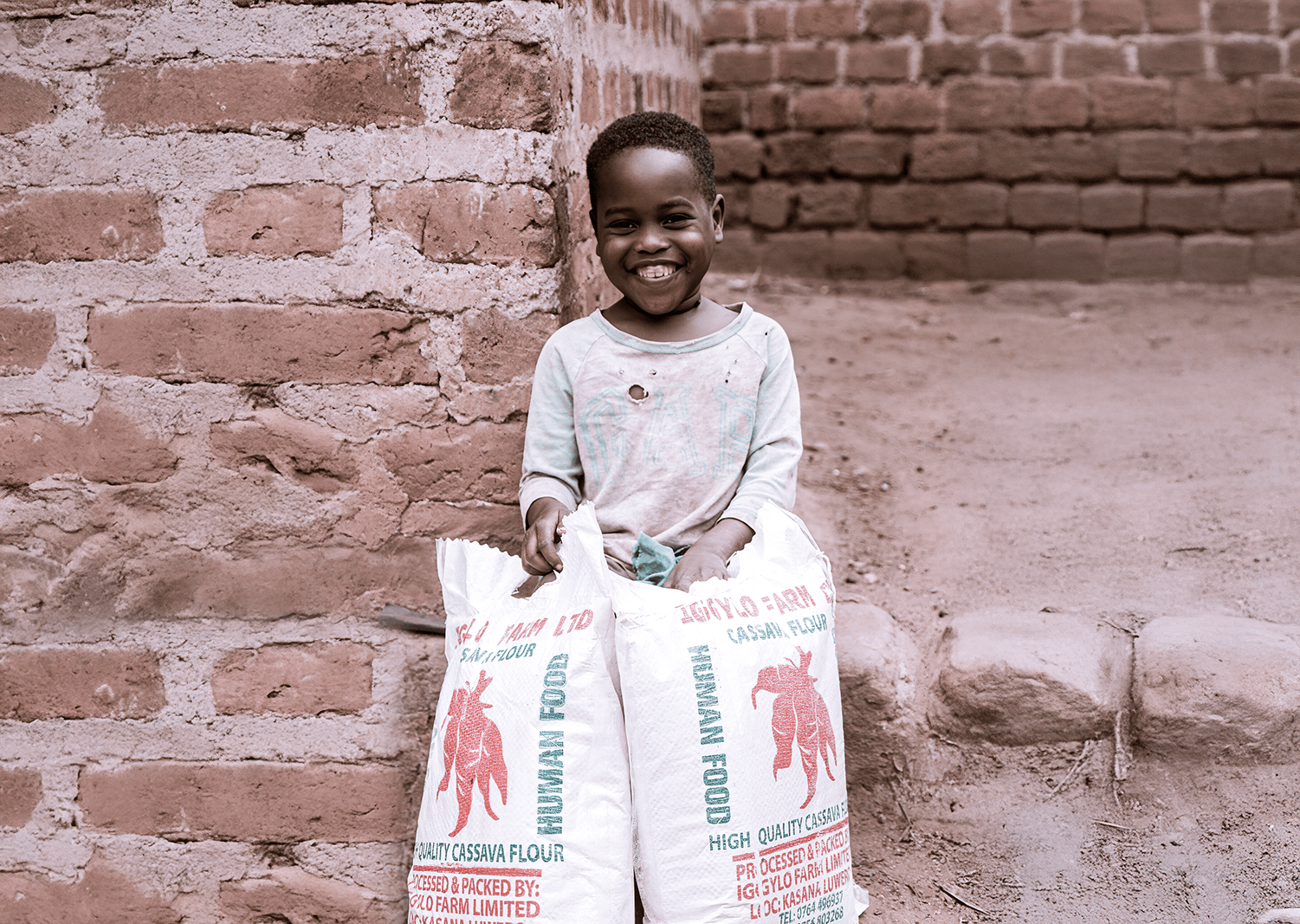 A girl kneels outdoors holding two bags of cassava flour near a brick wall.