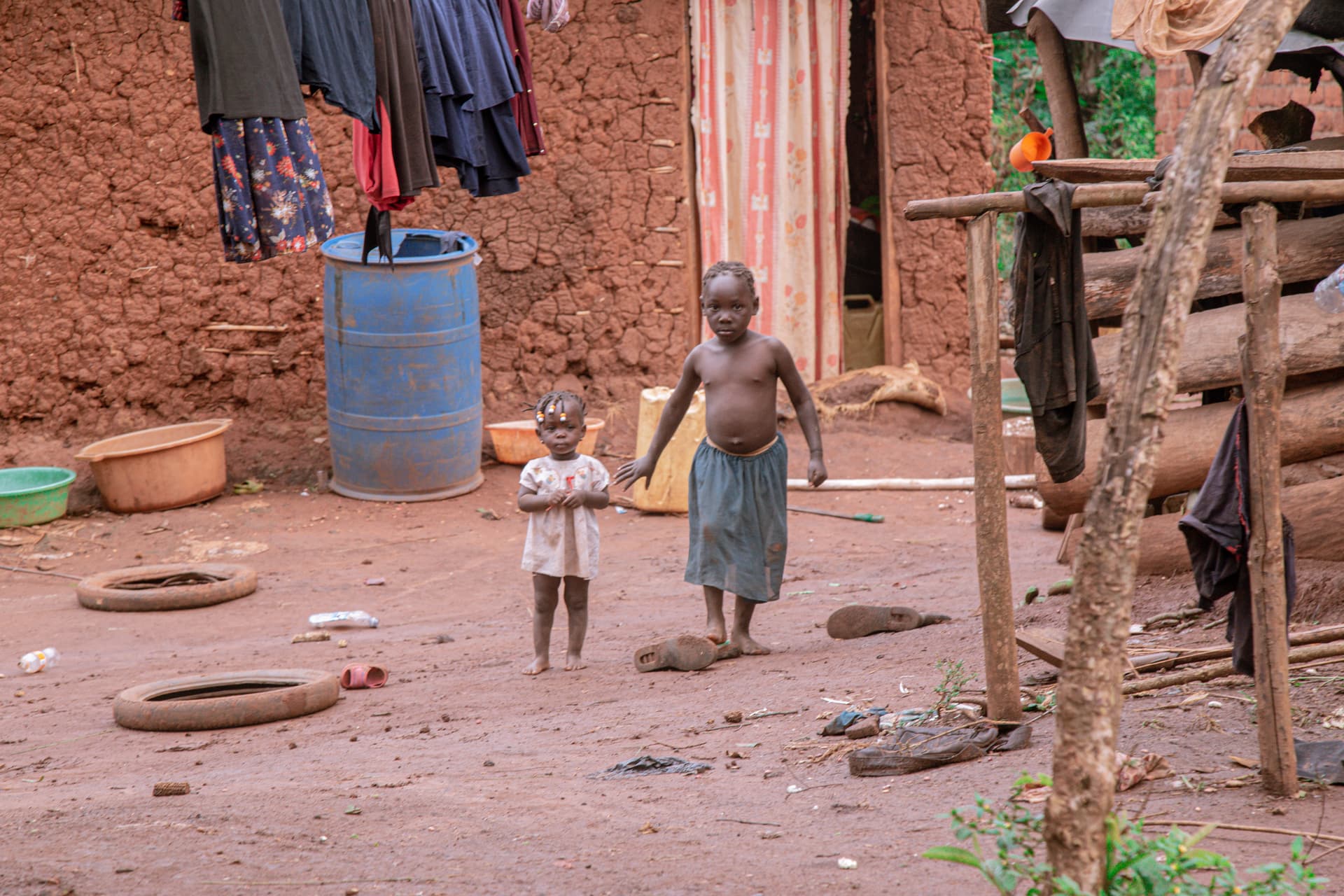 Wide view of a rural homestead with laundry drying, a blue barrel, and children in the yard.