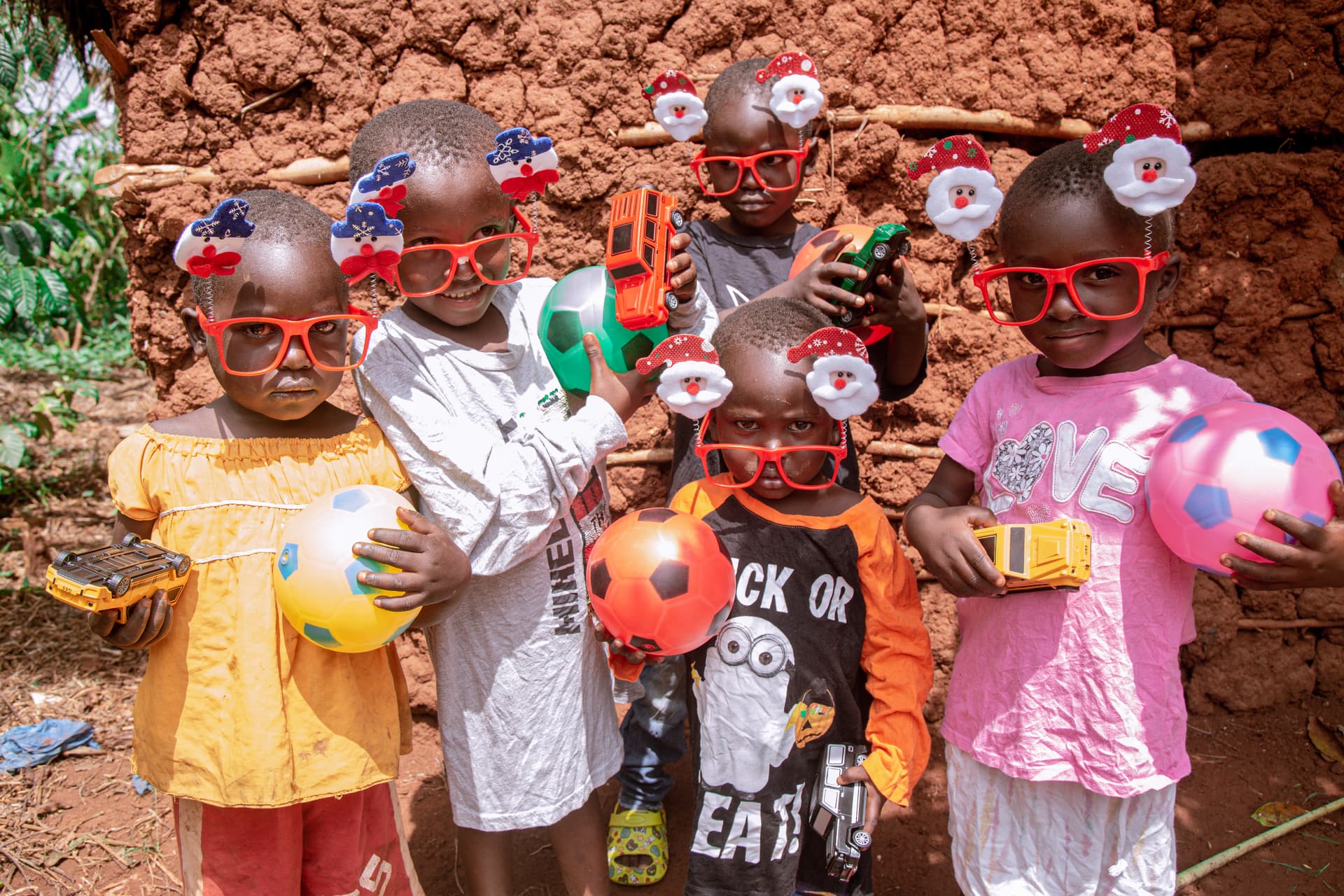 Five Ugandan children wearing red novelty glasses hold colorful balls in front of a mud brick wall.