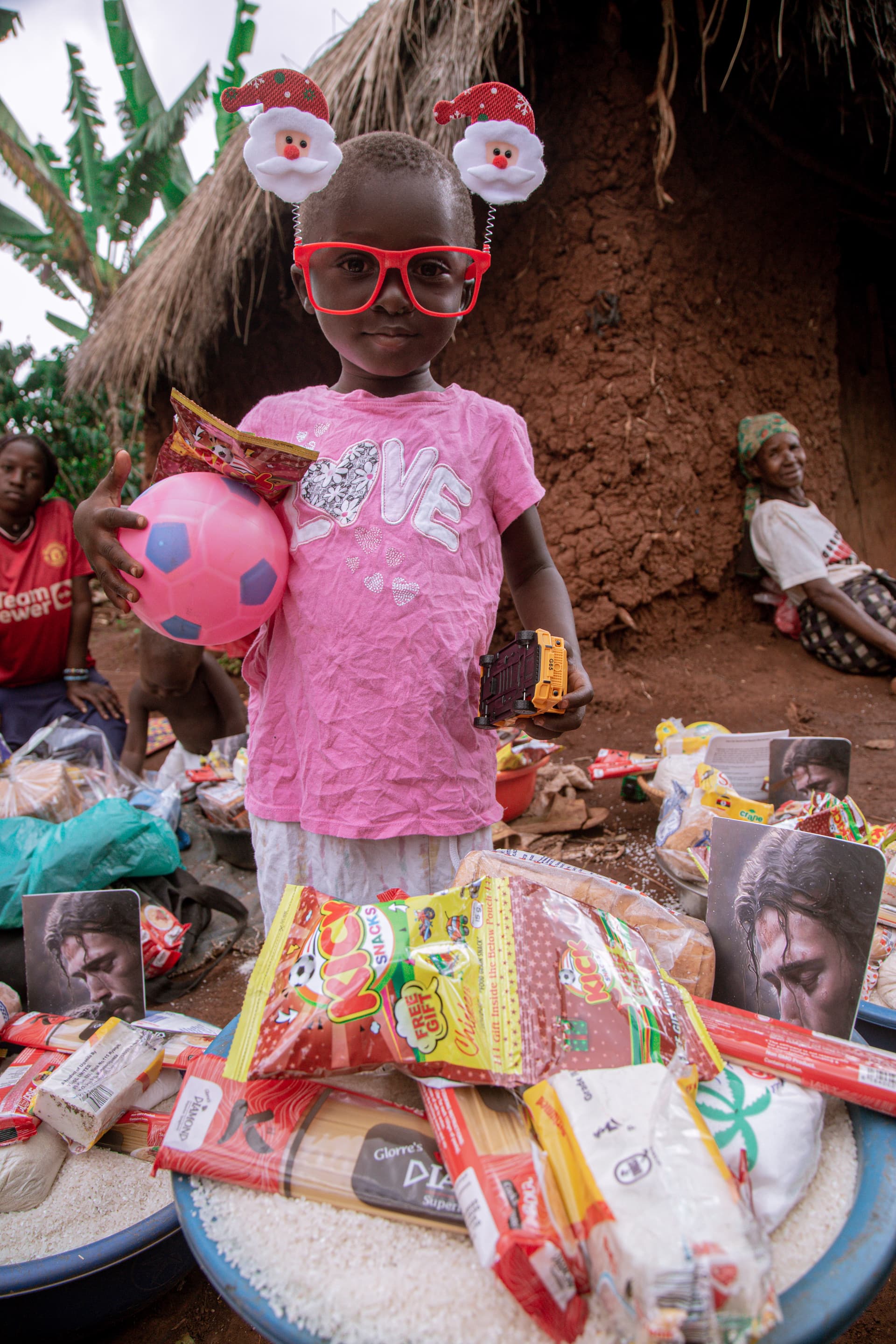 A girl in a pink shirt wearing a festive headband and sunglasses holds a pink ball beside a basin of gifts.