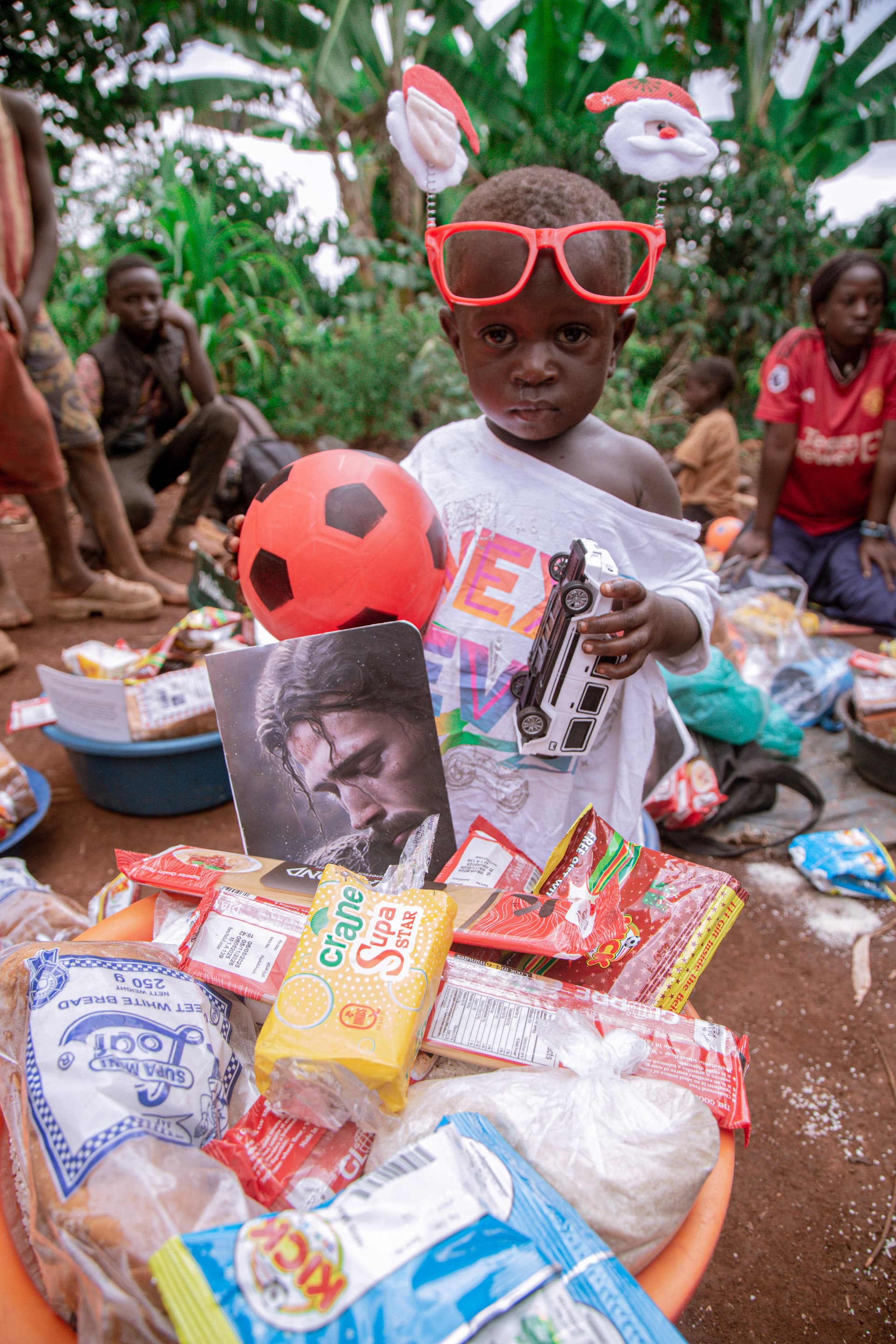 A boy wearing red sunglasses and a reindeer headband sits behind a basin of Christmas food packages.