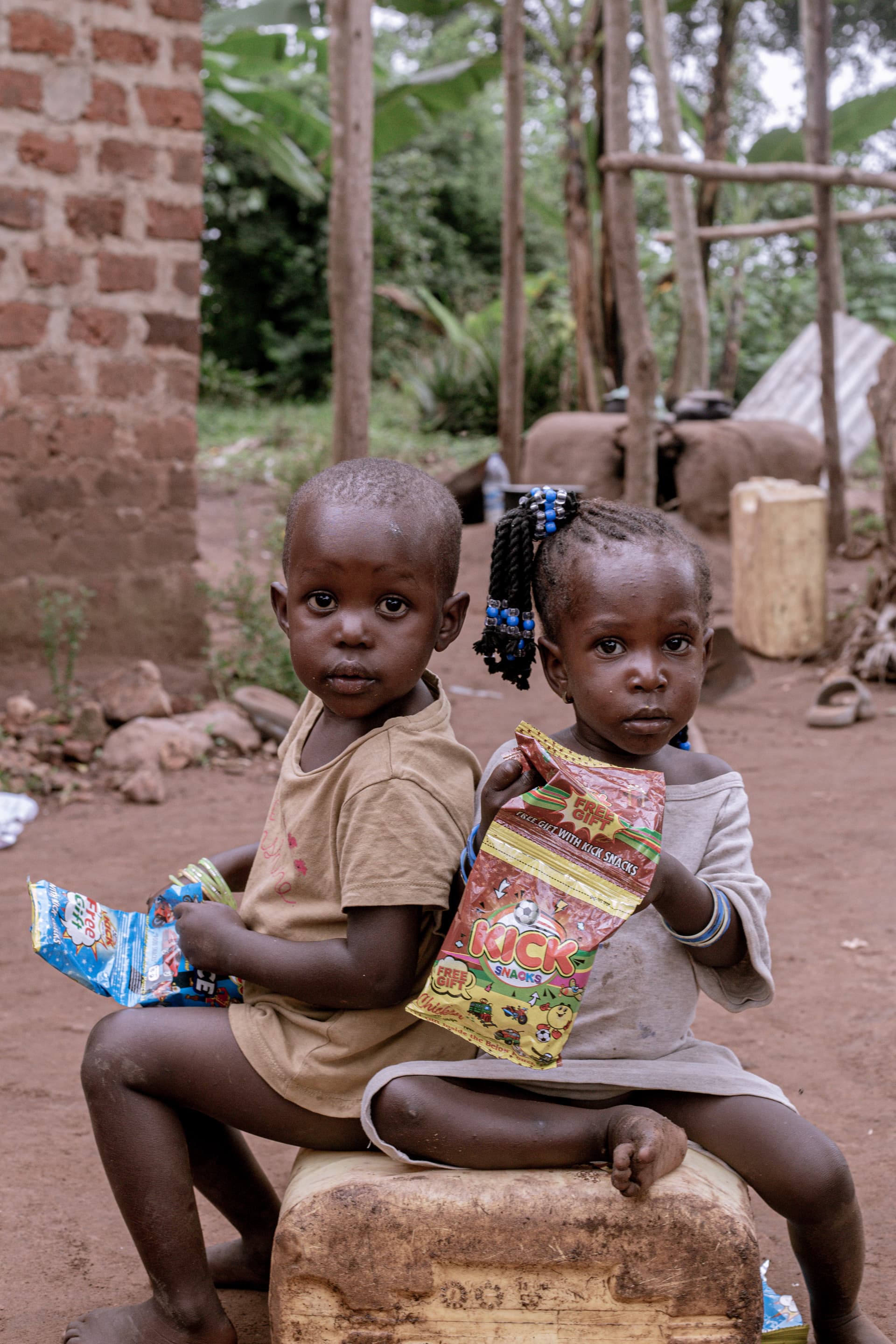 Two children sit together on a stone ledge holding colorful food packets from a distribution.