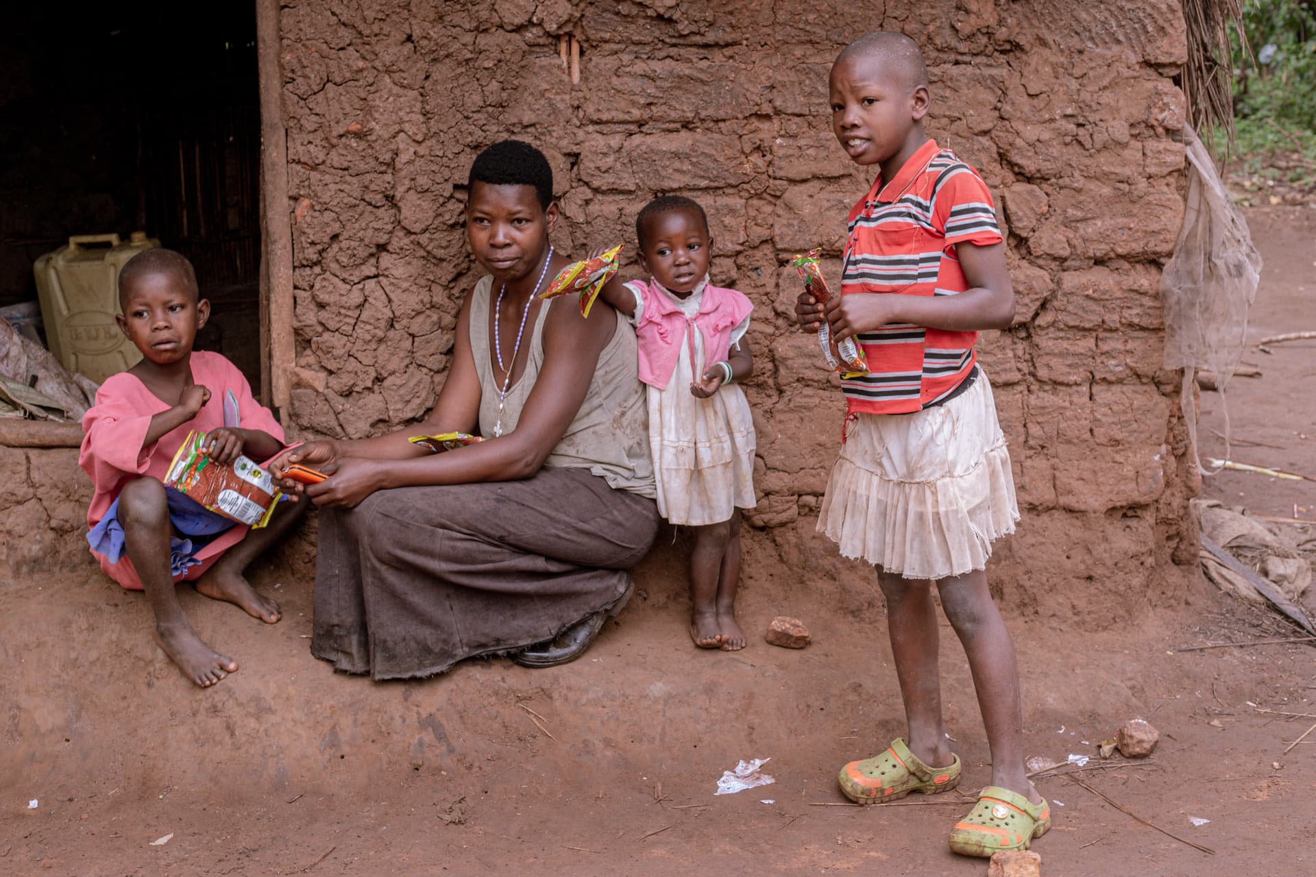 A woman sits outside a brick home with two children, one girl in pink holding food packets.