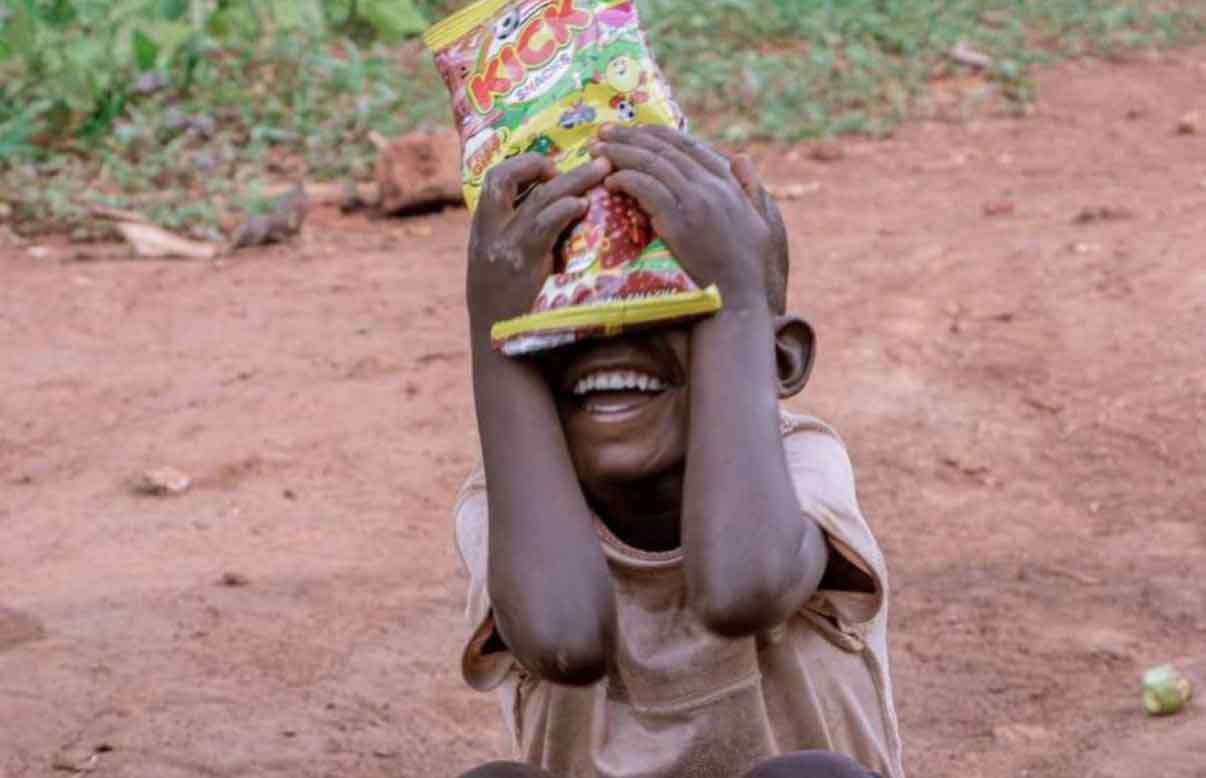 A boy laughs while holding a red and white patterned party hat over part of his face.