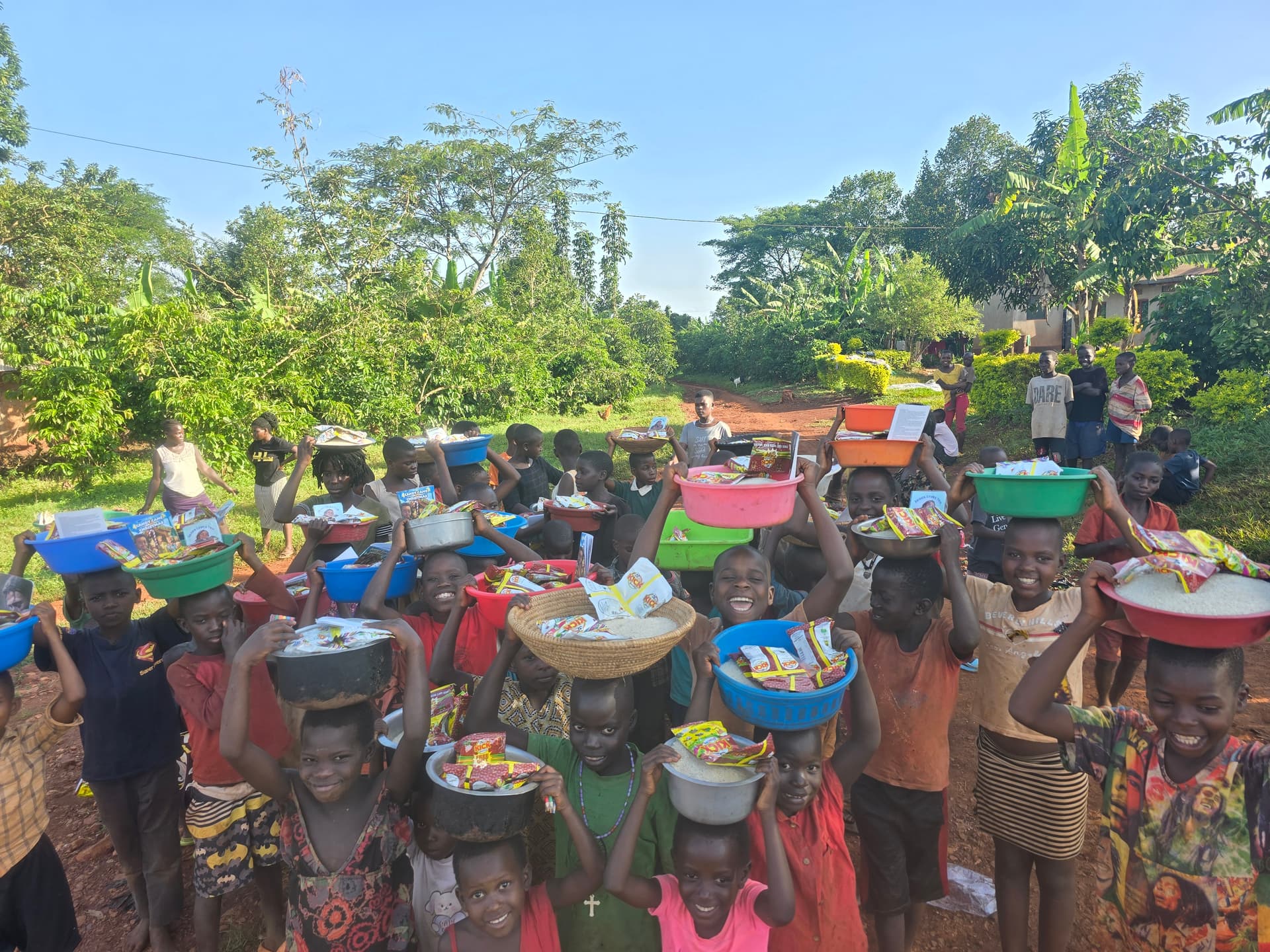 Line of children walking along a dirt road carrying basins of food on their heads.