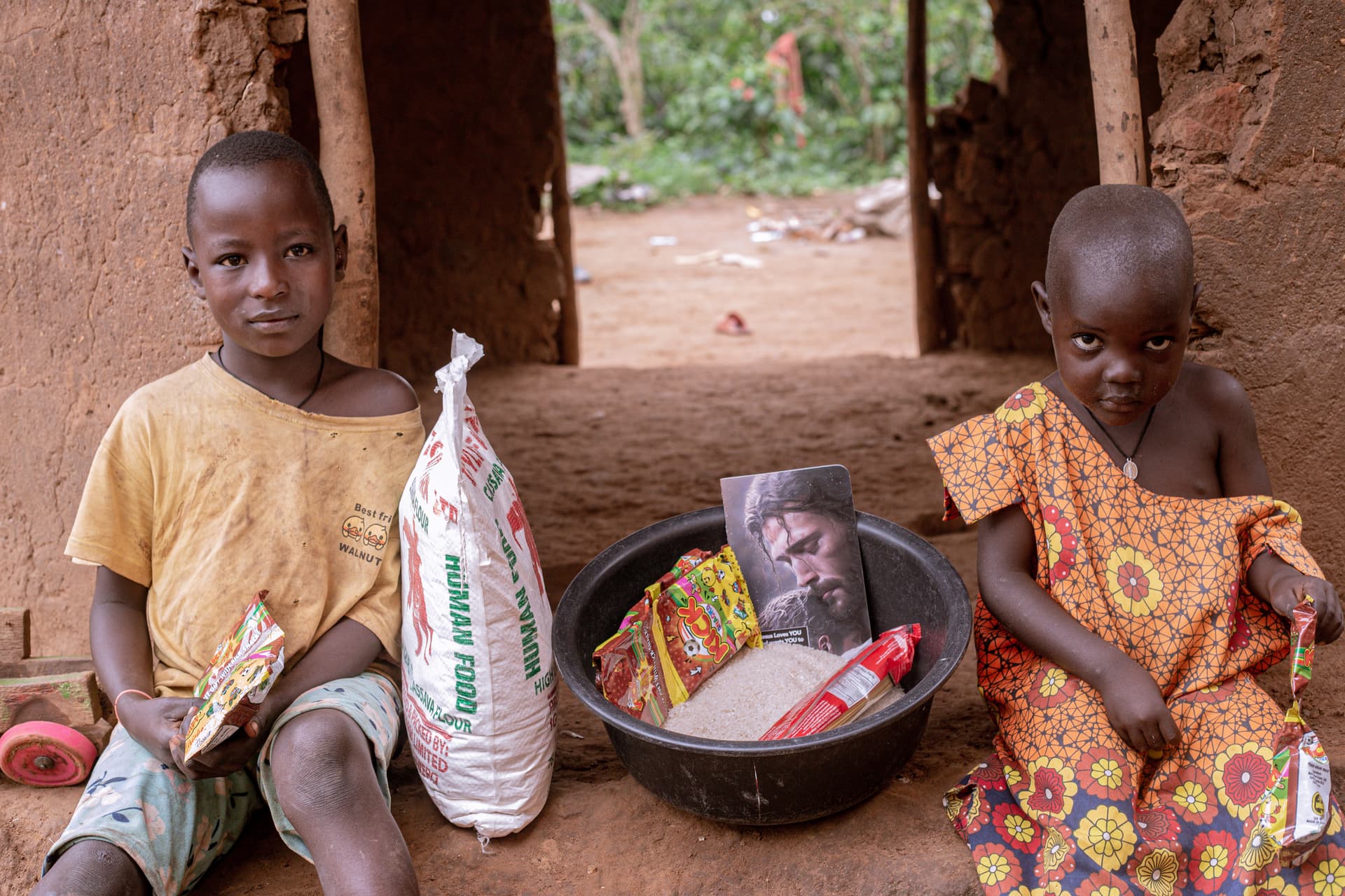 Two children sitting on a porch with a sack of rice and a bowl of food, one holding a gospel tract.