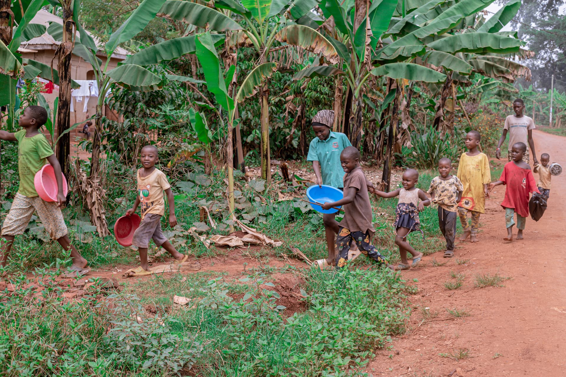 Children run and play along a dirt path lined with banana plants in the countryside.