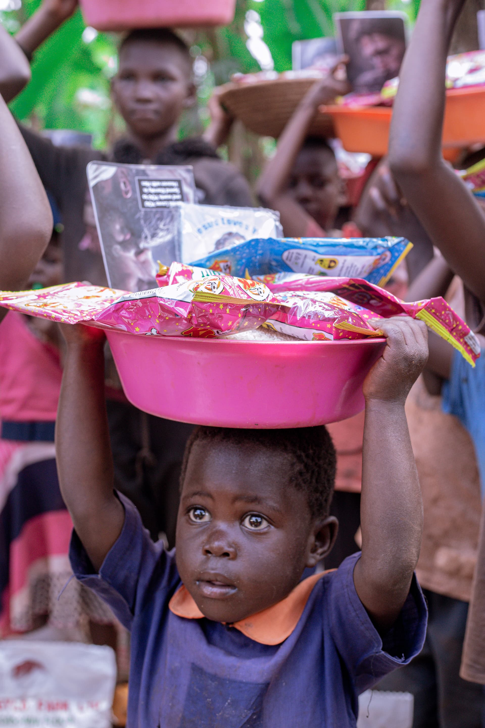 Children in a forest clearing carrying a pink basin filled with food packets on their heads.