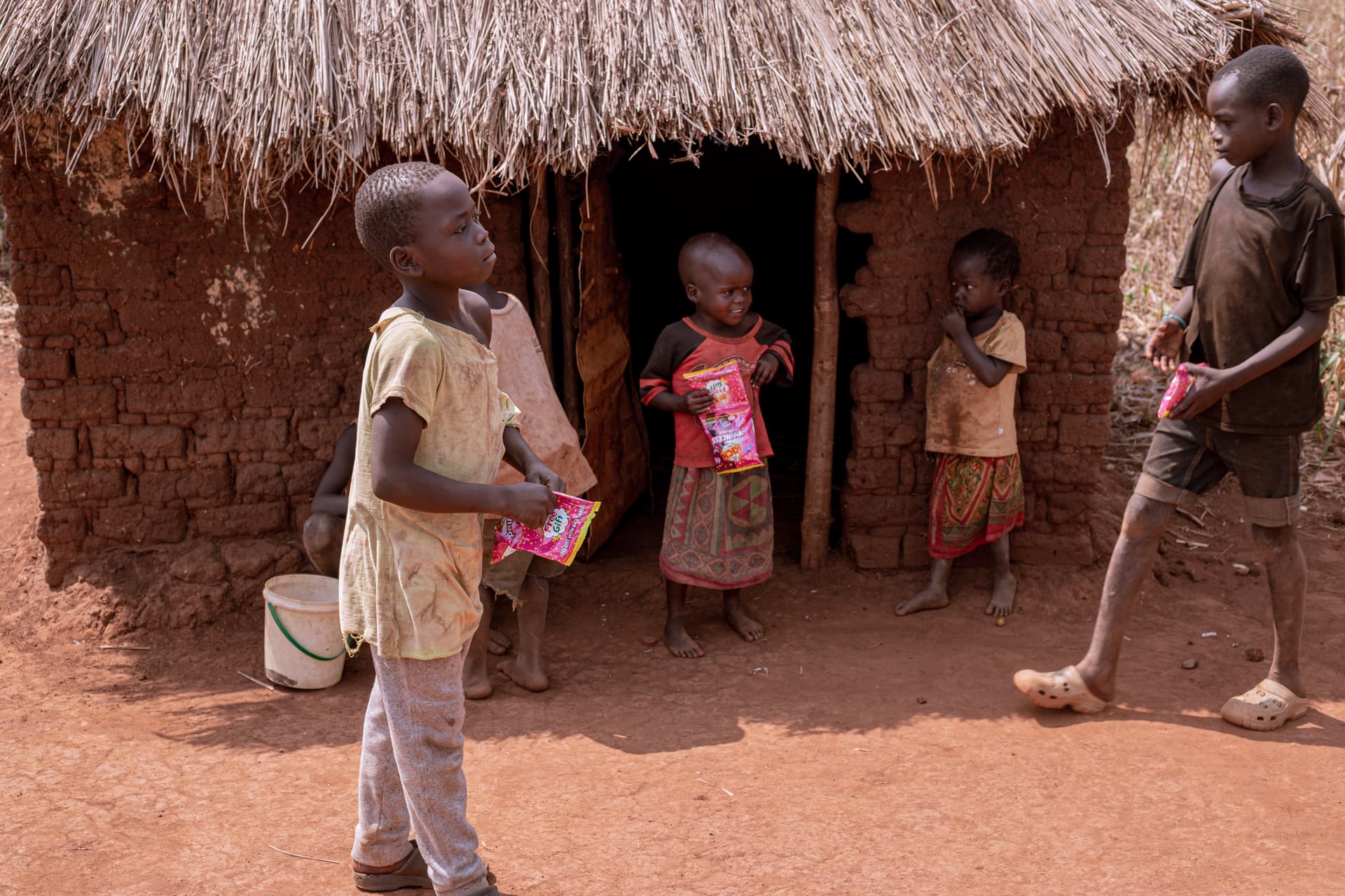 Children standing outside a mud hut, one holding a food package and another holding a stick.