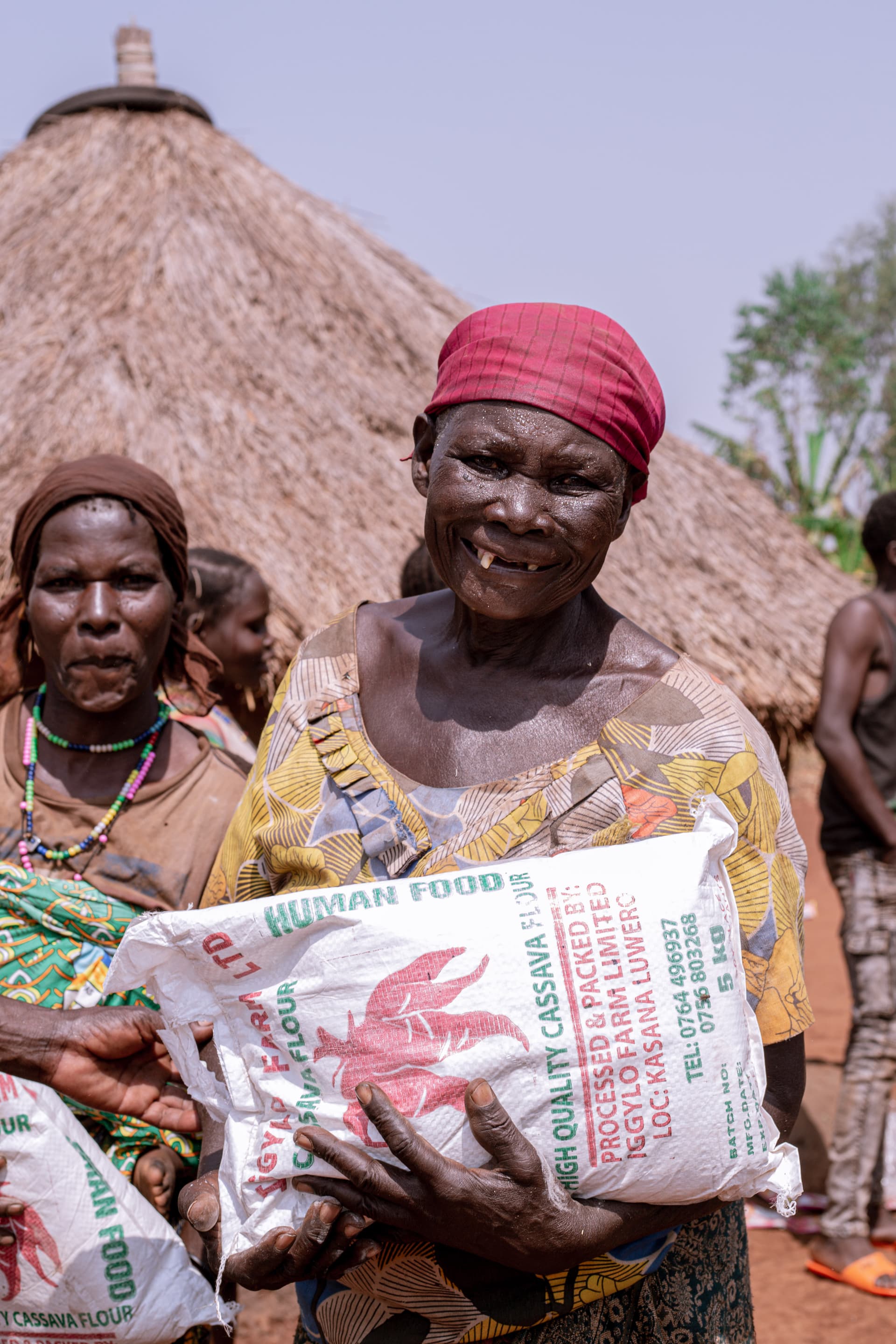 Elderly woman in a red headscarf holding a bag of food outside a thatched hut.
