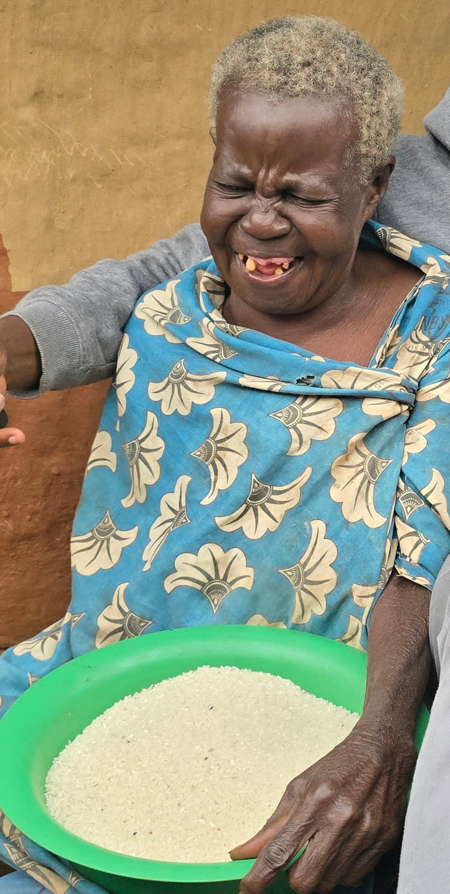 Elderly woman in a floral dress holding a green basin of grain outdoors.