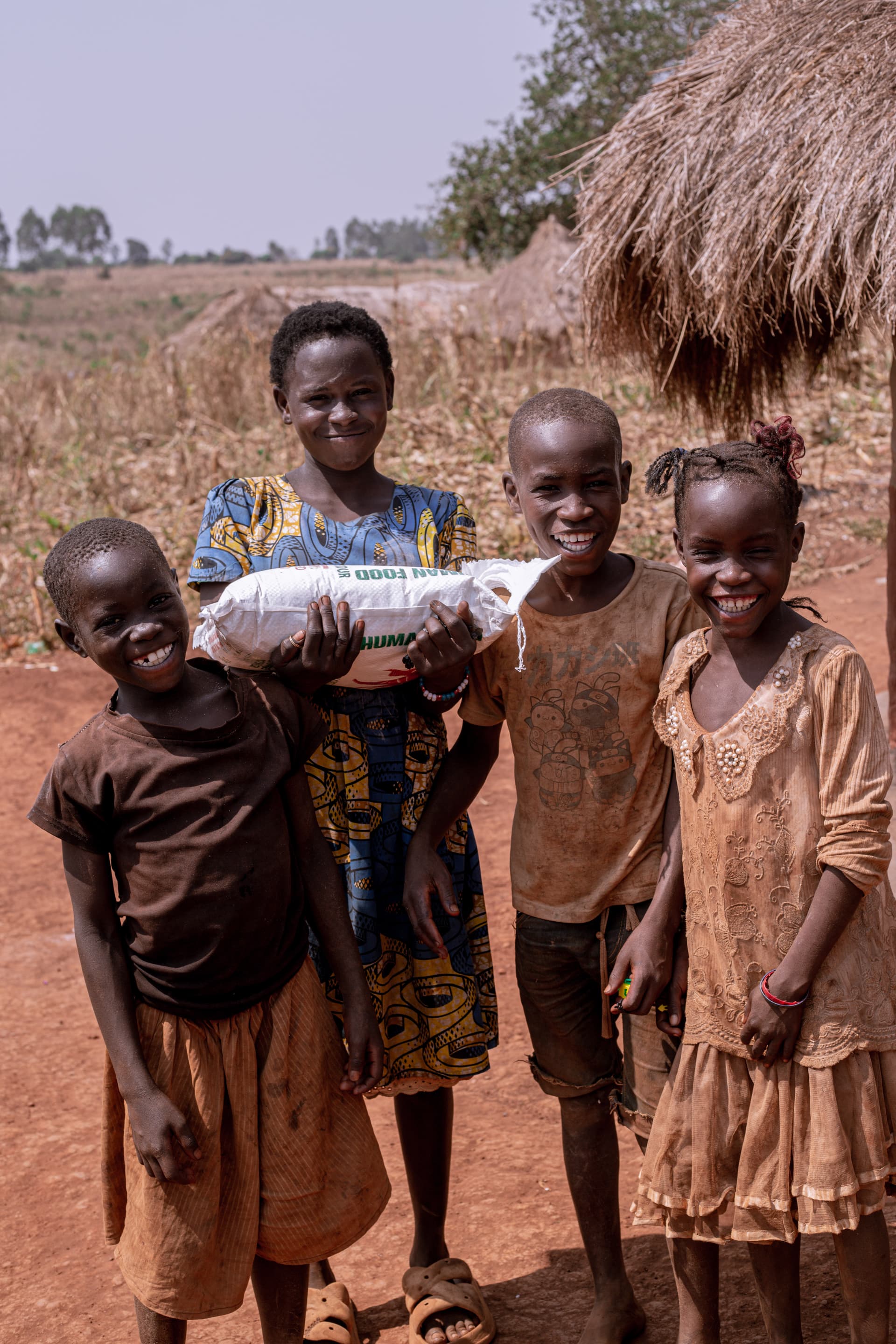 Woman holding a sack of food standing with children outside a thatched hut.