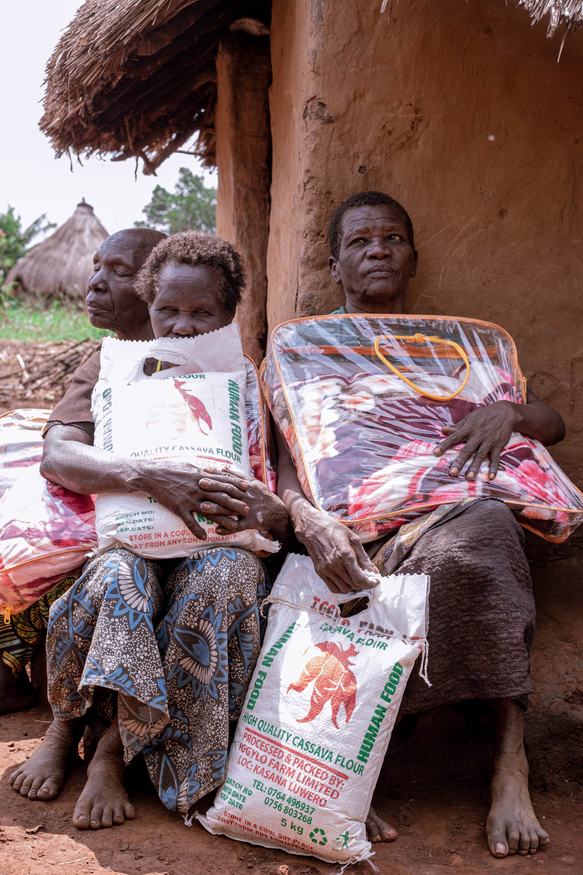 Elderly woman seated outside a mud hut holding a grandchild and a bag of food alongside a folded blanket.