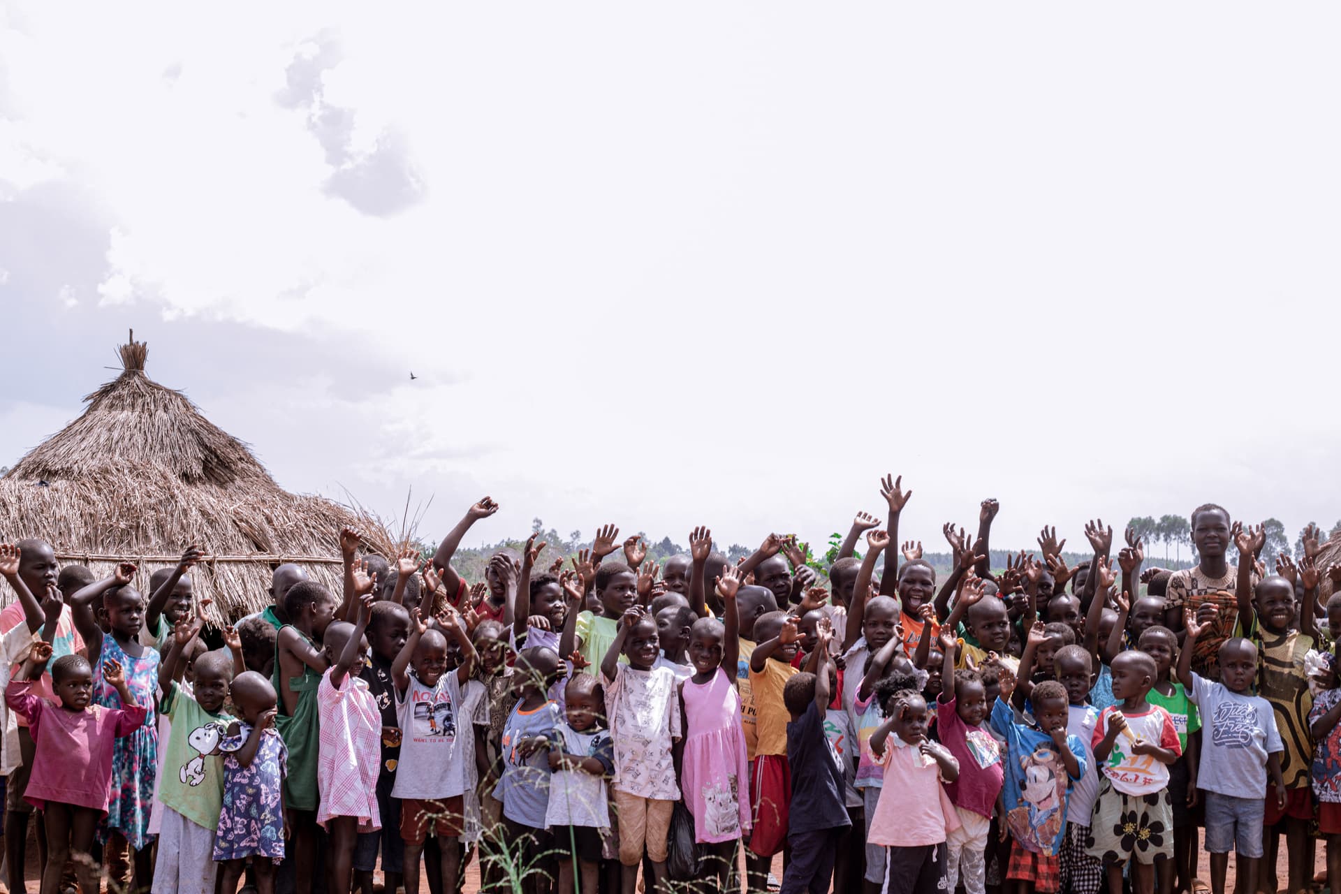 Large crowd of villagers gathered under thatched roofs, many raising hands during a JFM outreach event.