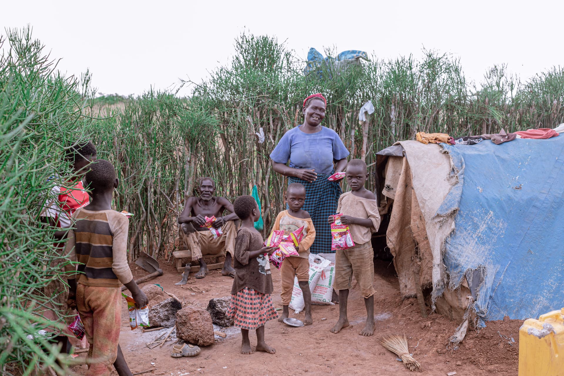 Woman and children standing with sacks of food beside a patched tarp shelter in a field.