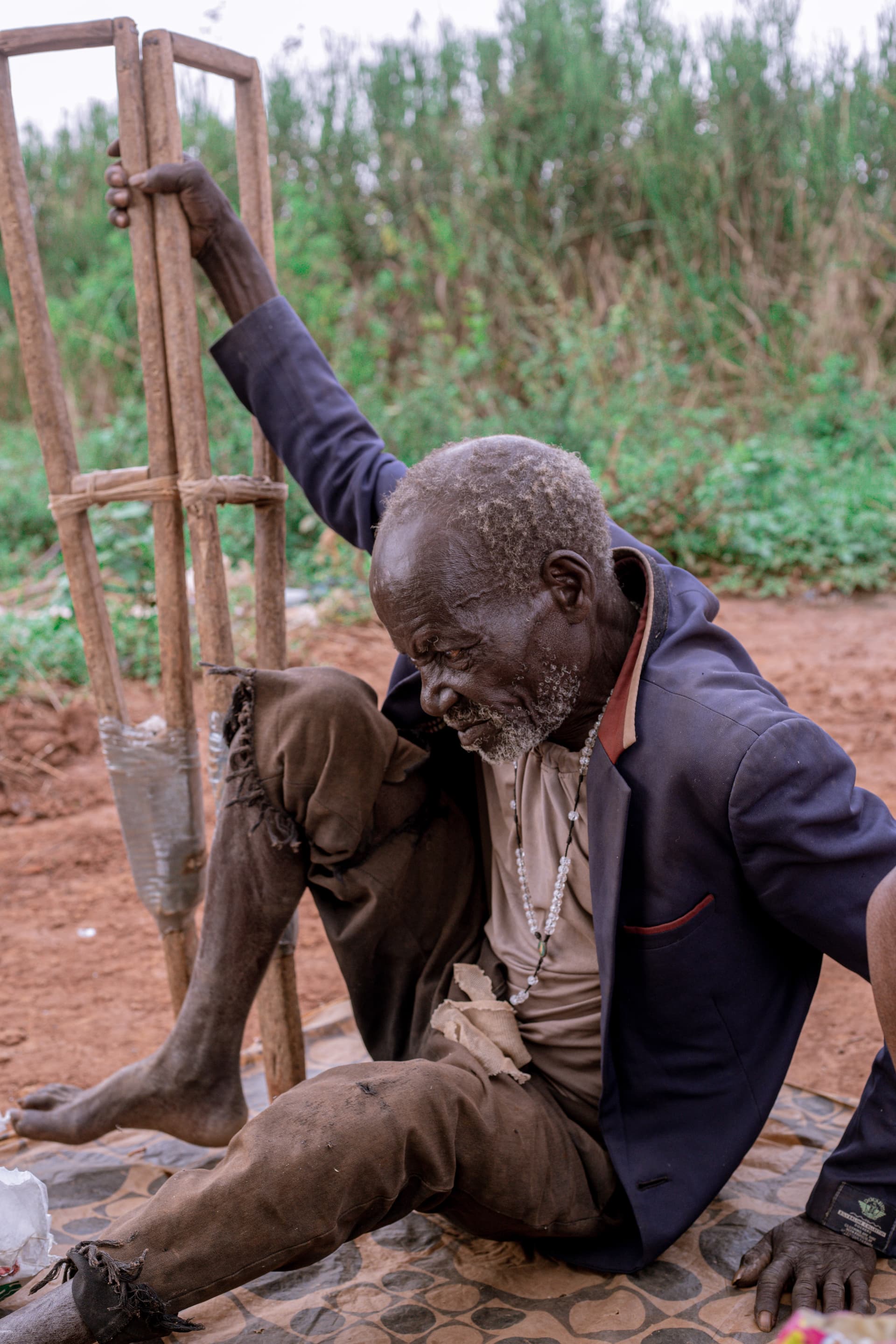 Elderly disabled man sits on the ground holding a wooden frame, reaching upward.