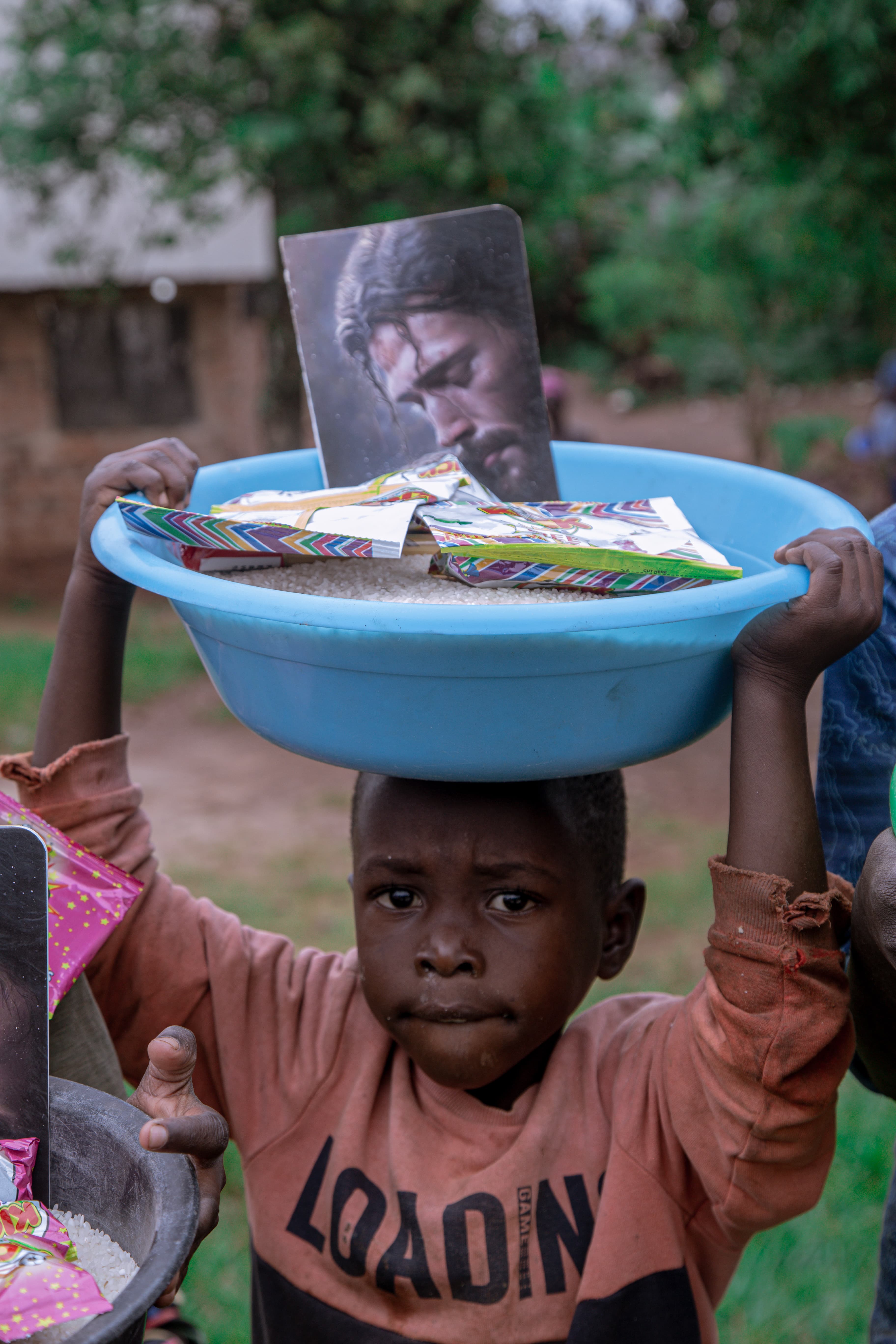 Boy balances a blue basin holding food items and a printed photo on his head.