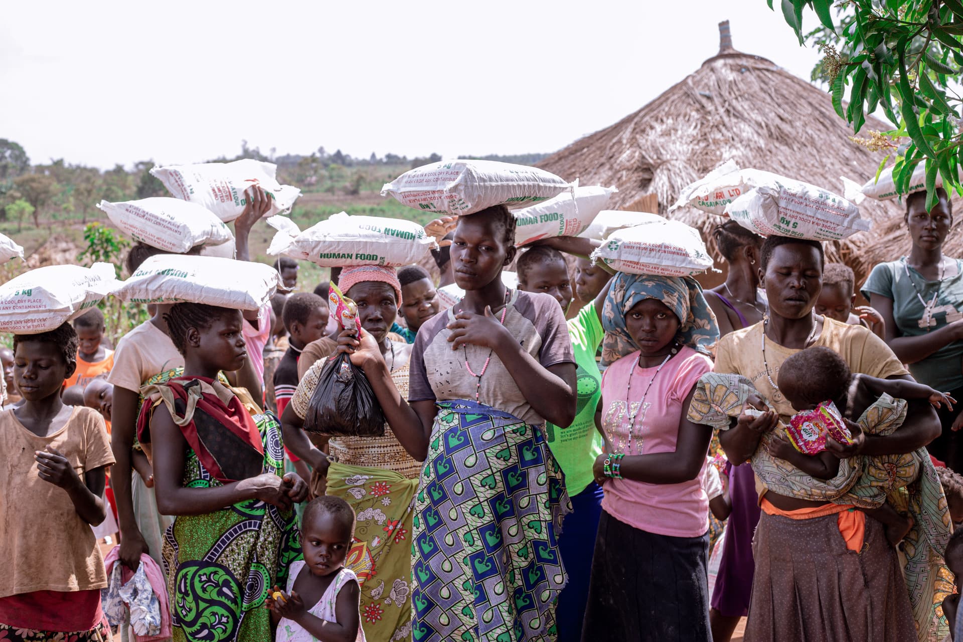 Line of women and children carry white food sacks balanced on their heads.