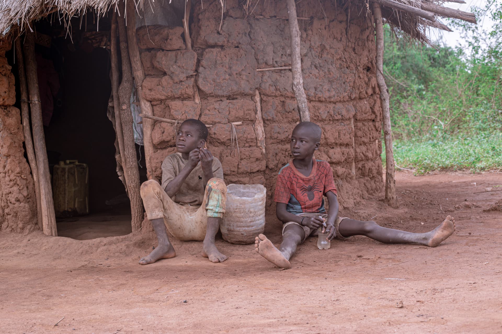 Two boys sit on the ground outside a mud-brick hut beside woven baskets.
