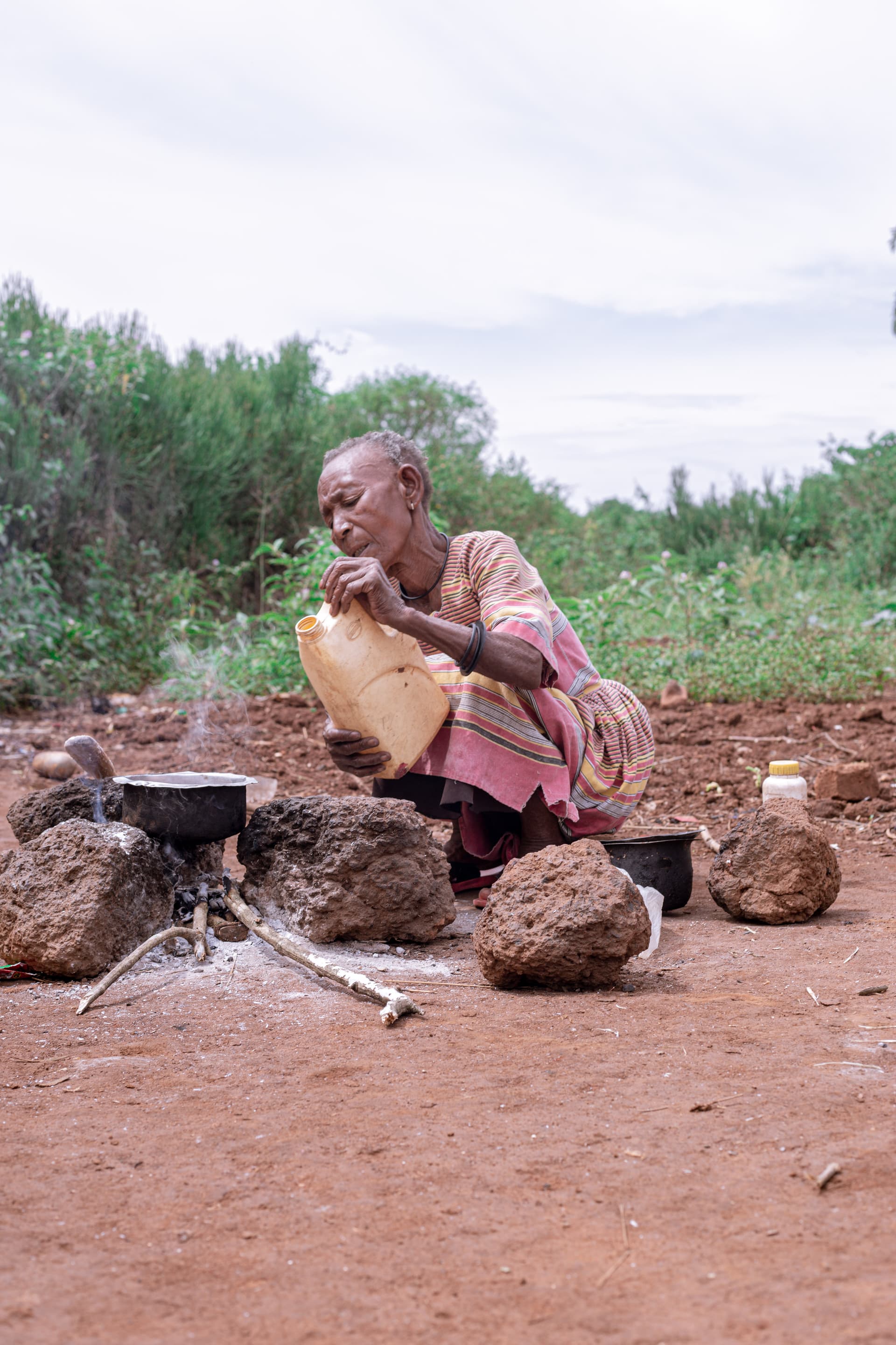 Elderly woman sits on the ground cooking over a small stove, sacks of goods nearby.