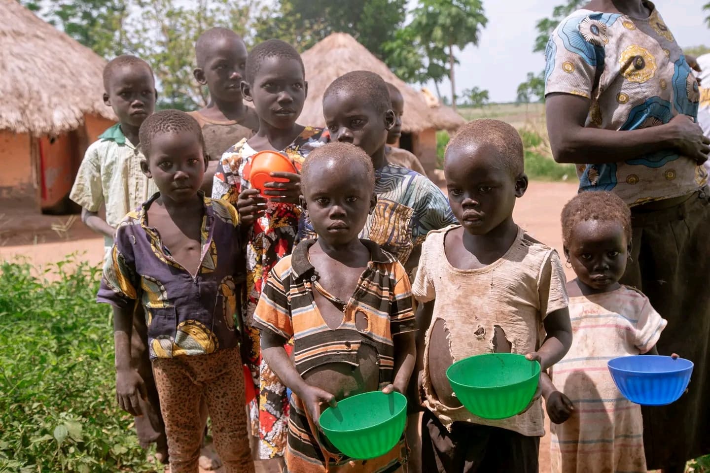 Ugandan children stand outside thatched huts holding green and blue plastic wash basins.