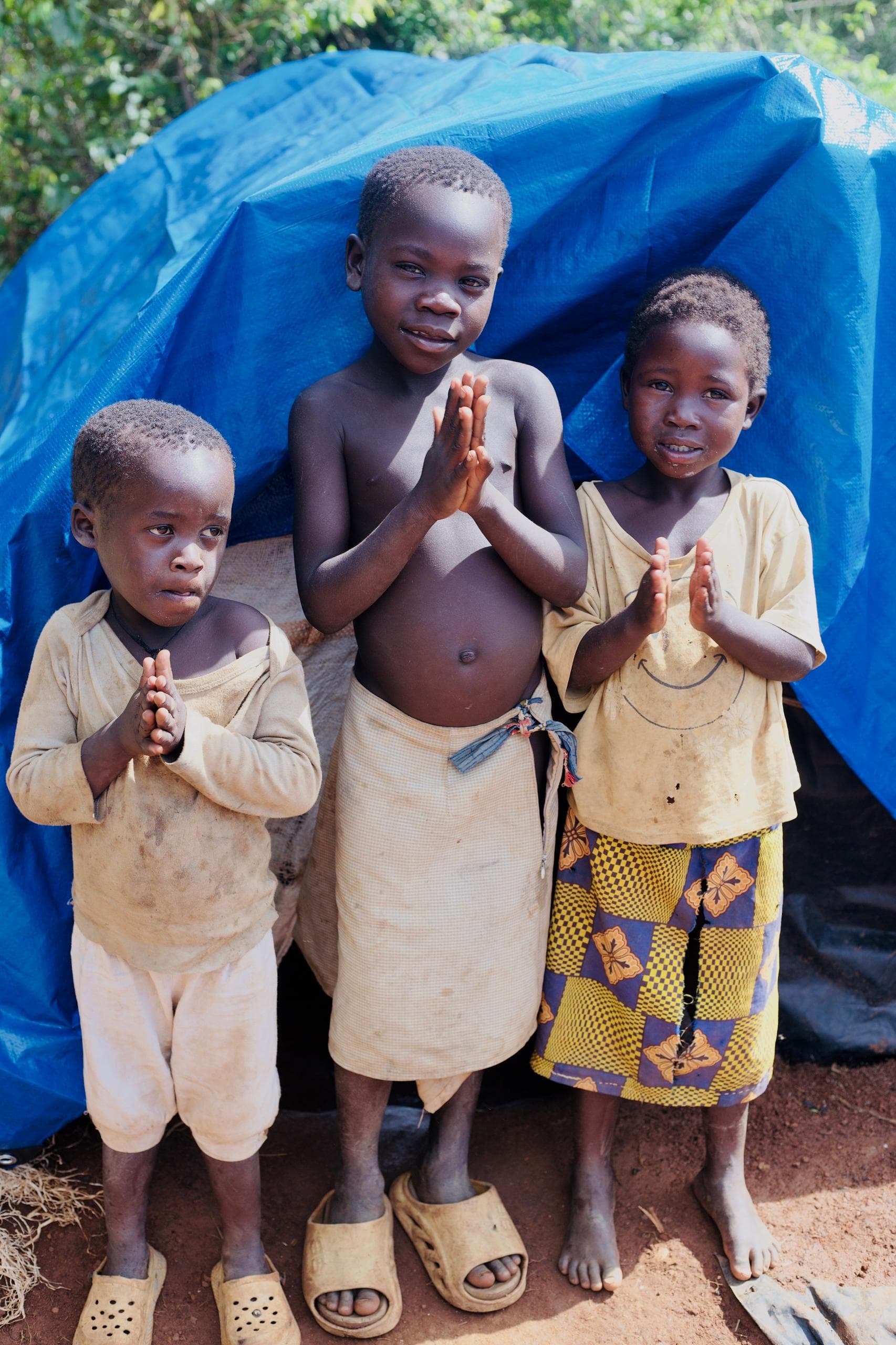 Three children with hands folded in prayer stand outside a mud-brick home.