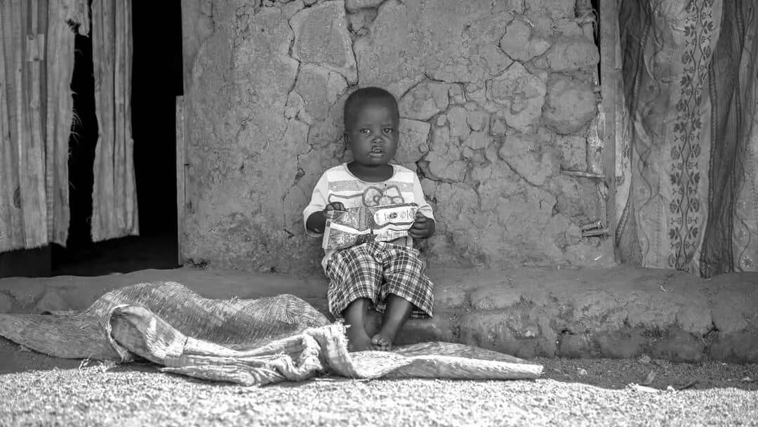 Black-and-white photo of a child sitting on a woven mat outside a hut.