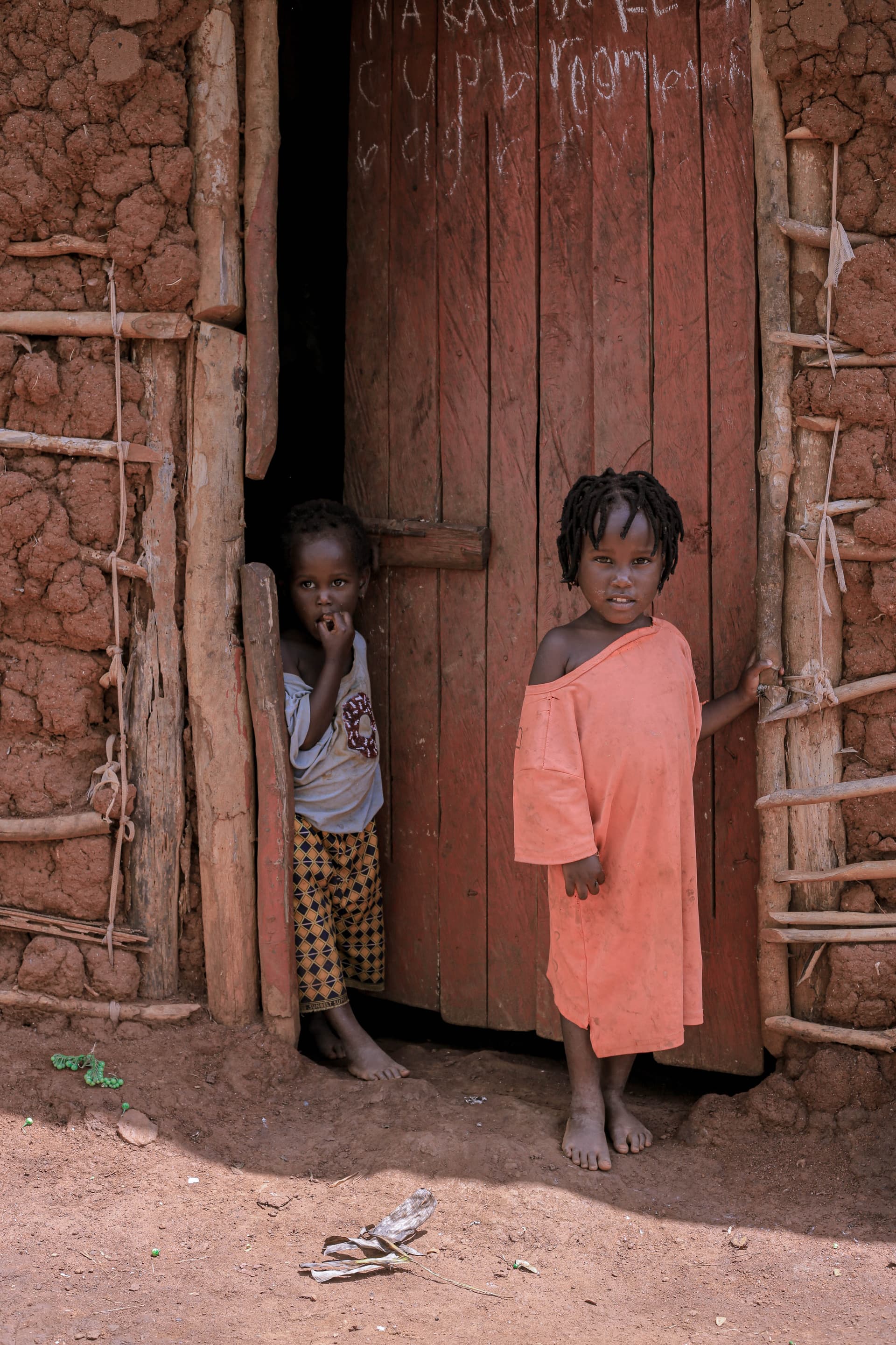 Two girls in a doorway of a wooden and mud hut, one wearing an orange dress.