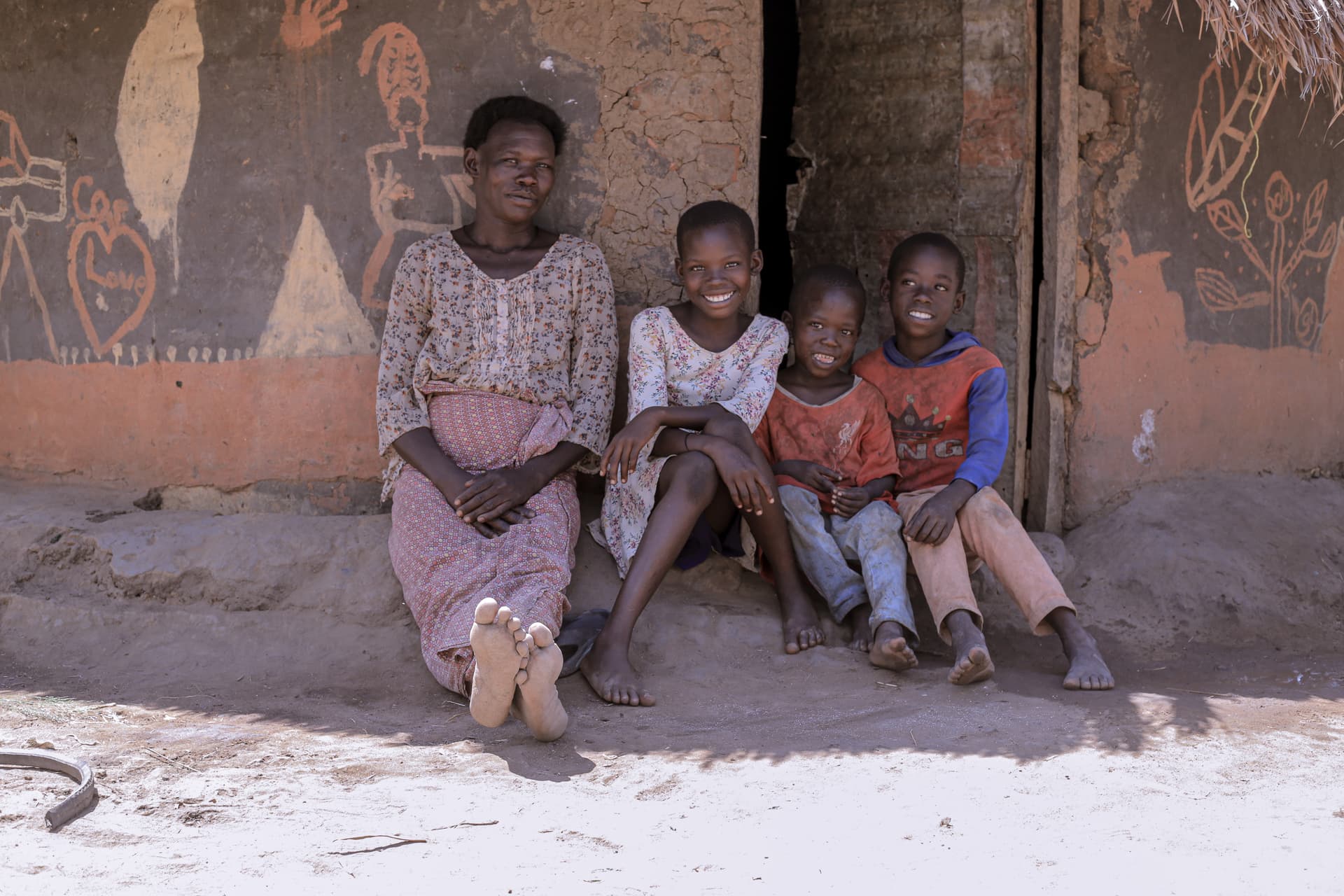 A family group sitting together on a bench outside a mud home painted with decorative patterns.