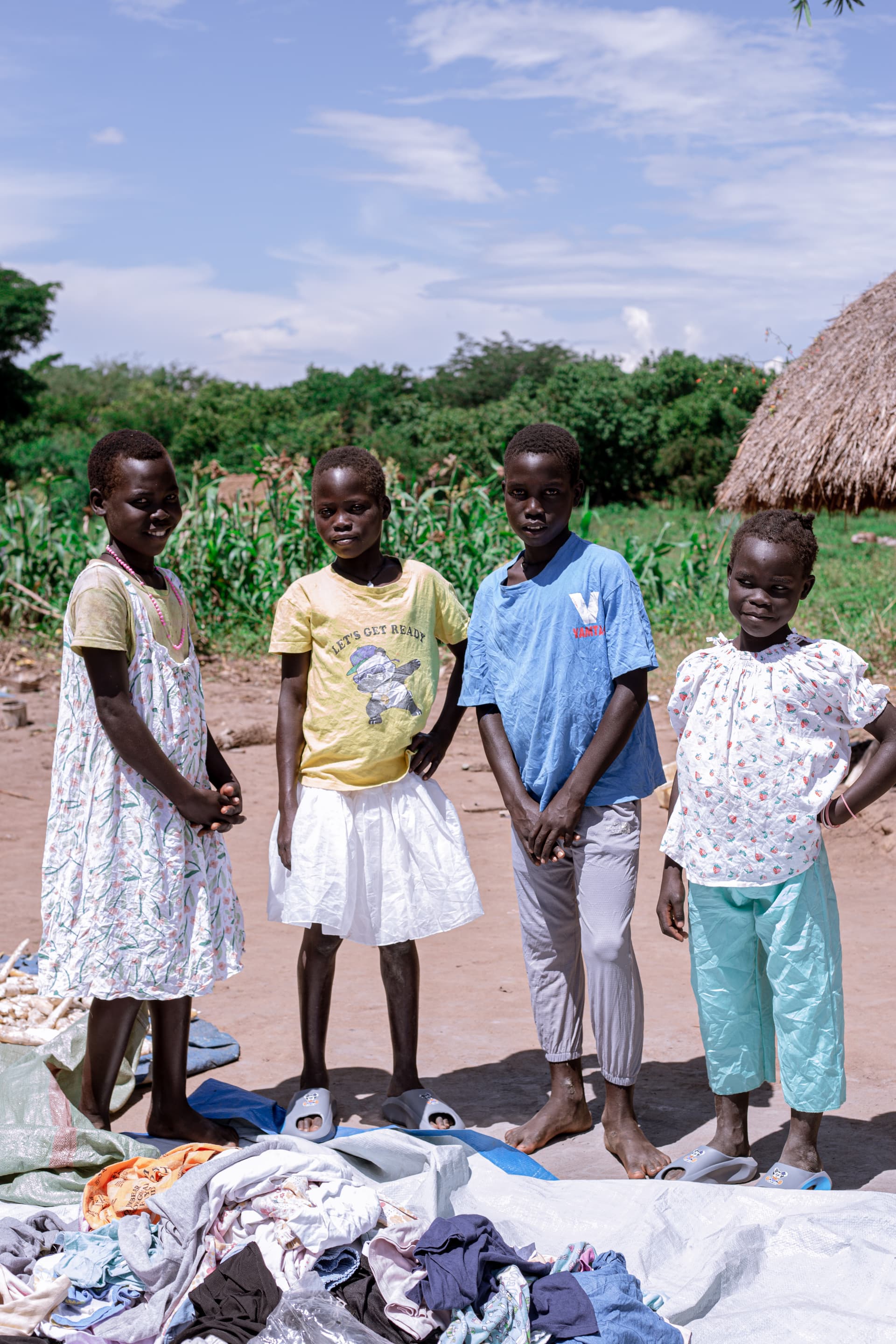 A group of children standing outdoors beside piles of donated clothing on the ground.