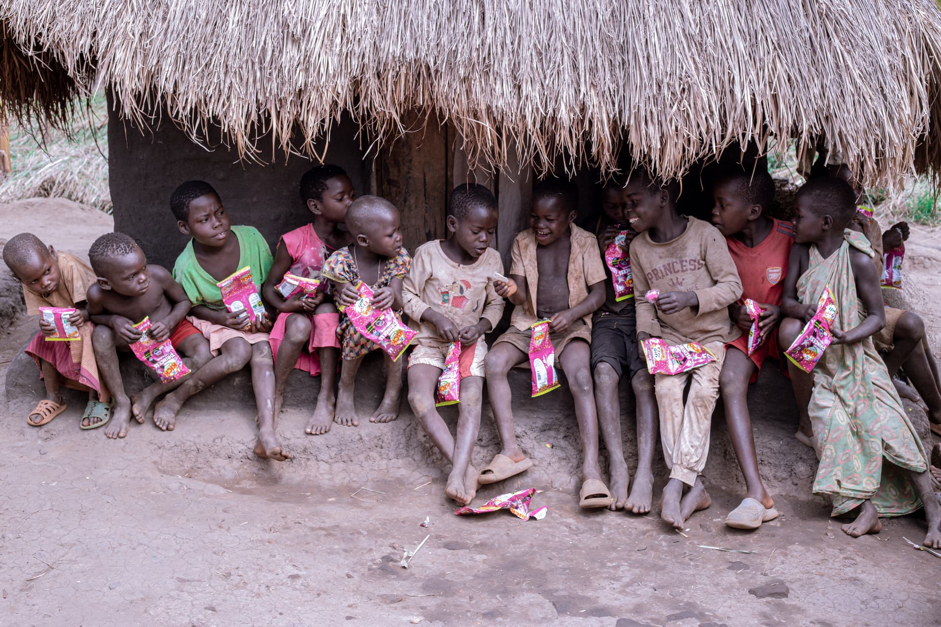 A row of children sitting under a thatched shelter each holding new clothing bundles.