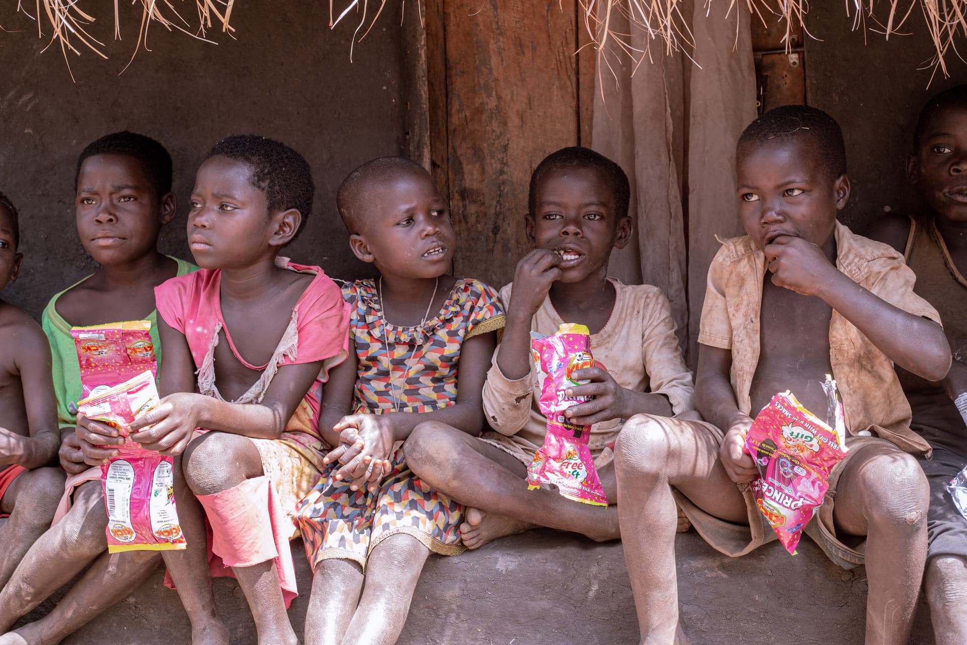 Several Ugandan children sitting in a row against a thatched hut, holding small food packets.