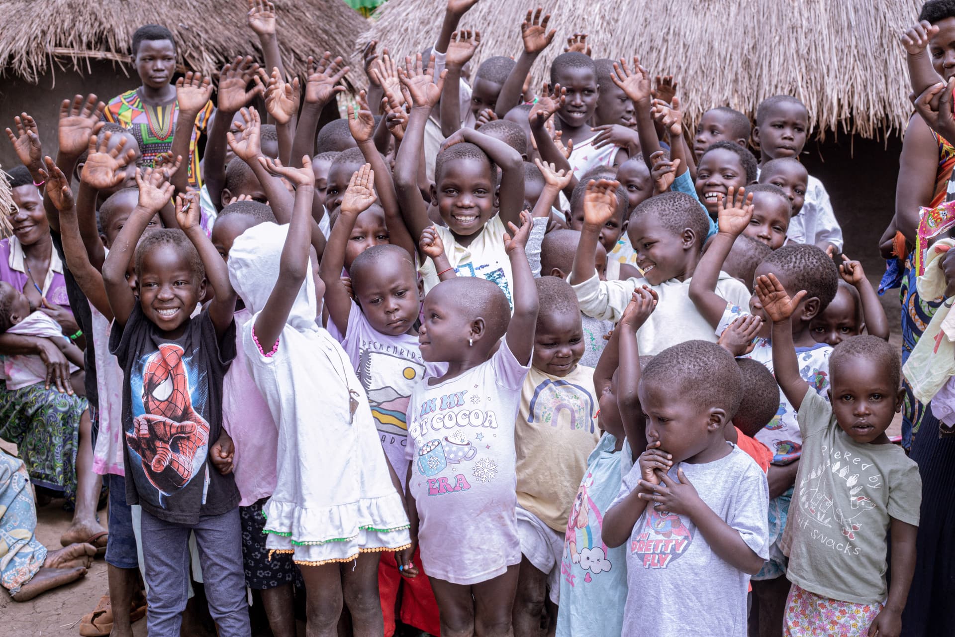 A large crowd of children and adults raises their hands and cheers outside a thatched building.
