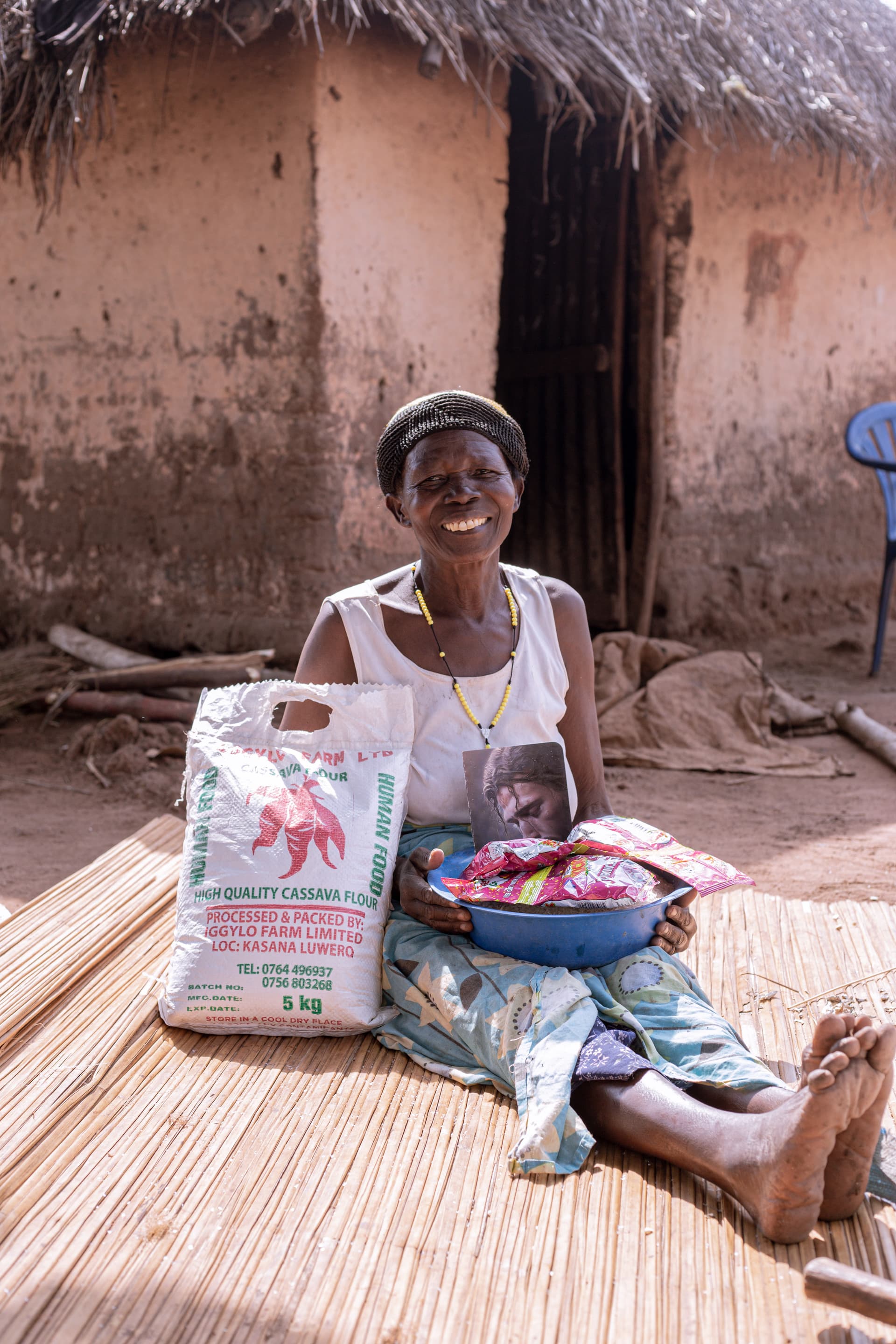 A woman sits on a mat outside a mud house beside a food package and basin.