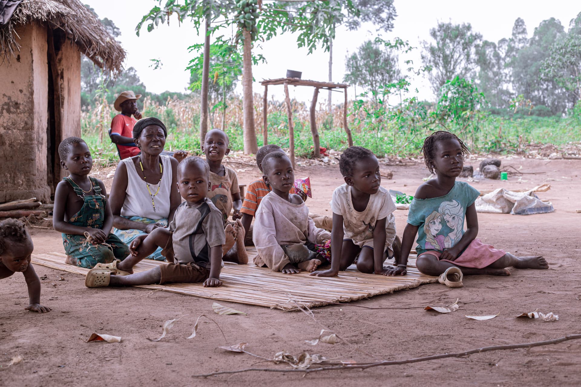 A group of children sits on a woven mat outside a mud house.