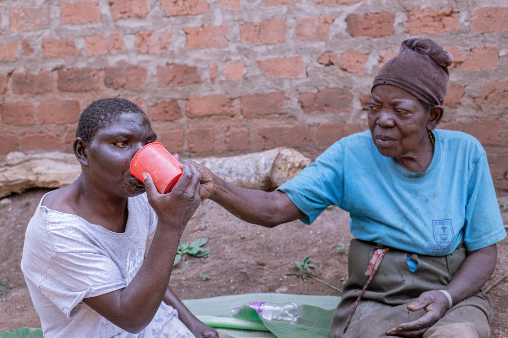 A woman feeds a person a drink from a red cup as both sit on banana leaves on the ground.