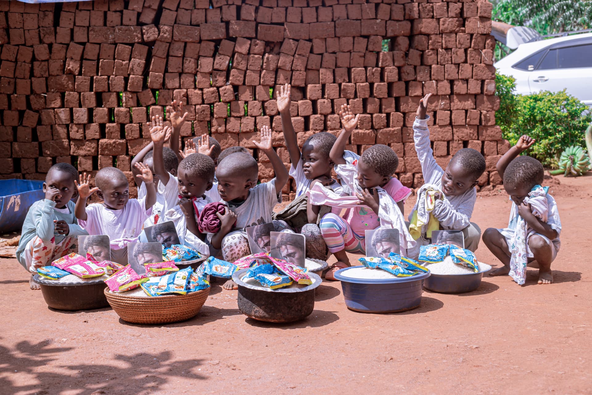 A row of children sit on the ground outside a brick wall, each holding a basin of food gifts.