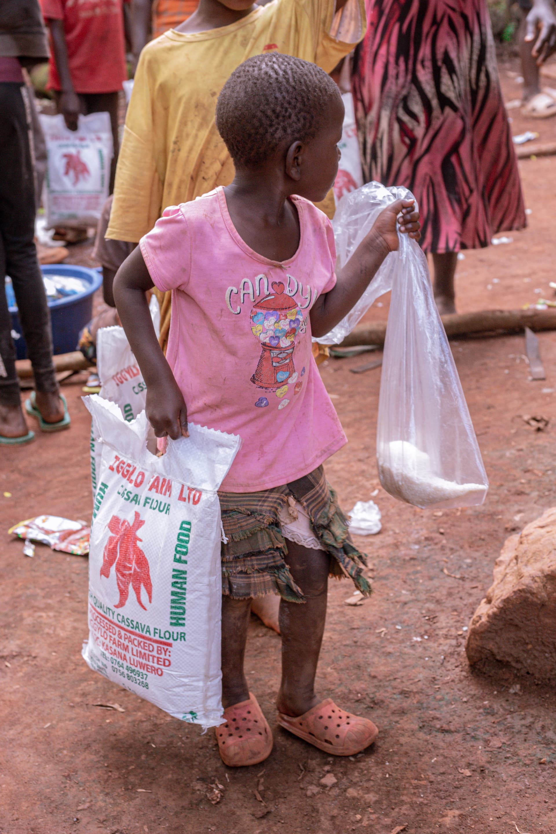 A girl in a pink shirt carries a plastic bag of food and a woven sack outdoors.